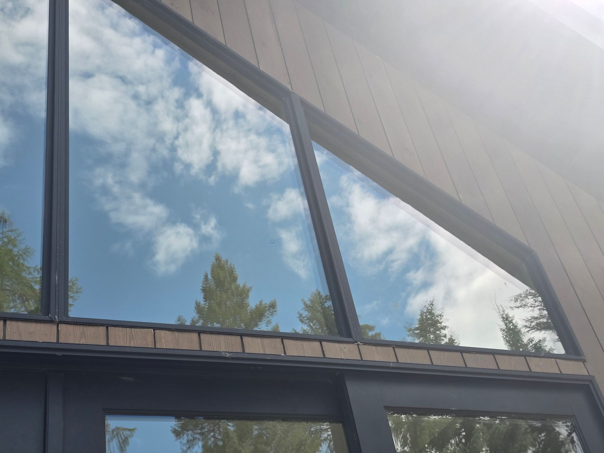 A low-angle view of a modern building with large glass windows reflecting a blue sky with clouds and surrounding trees.