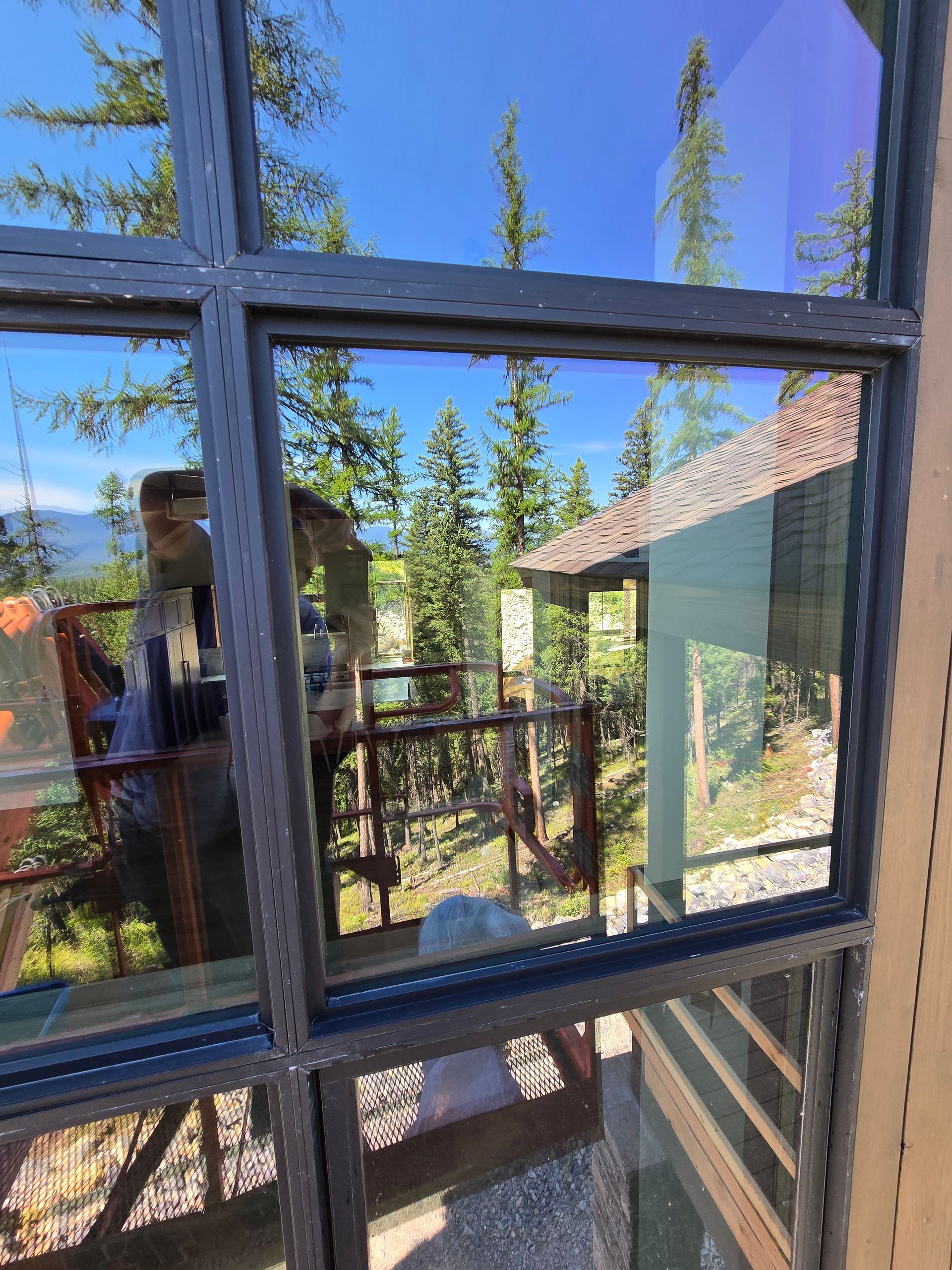 A window view looking out onto a forest and wooden deck on a sunny day, with a reflection of a person visible in the glass.