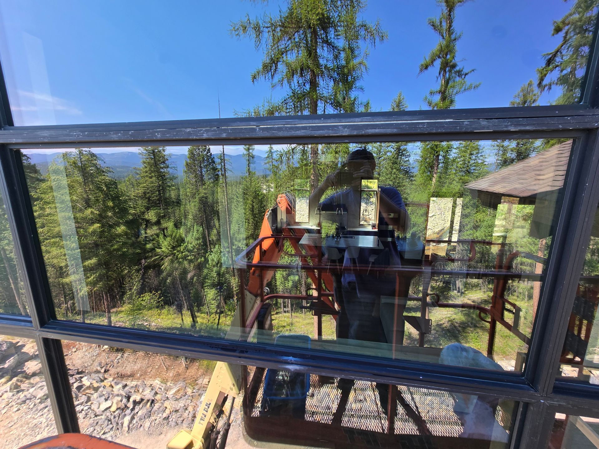 A person cleaning a large window, with a view of a forested mountain landscape and an orange excavator reflected outside.