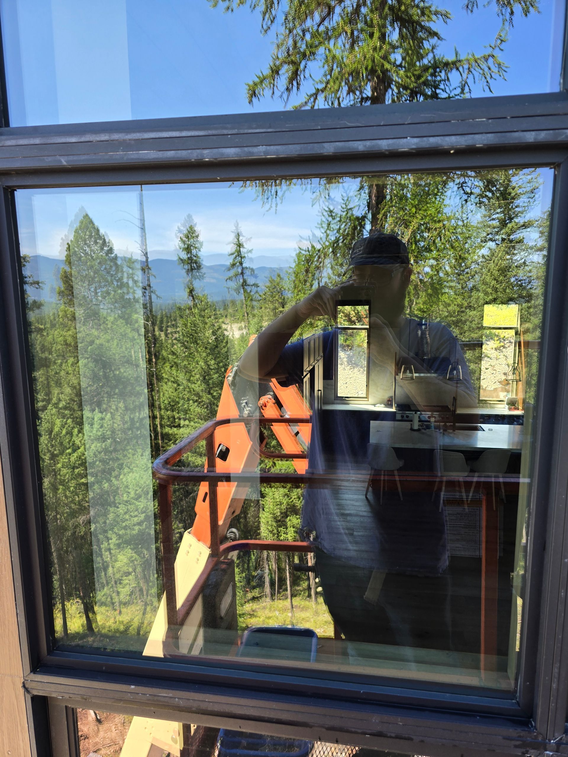 A person taking a selfie in a window reflection, showing a view of a forested mountain landscape and an orange crane.