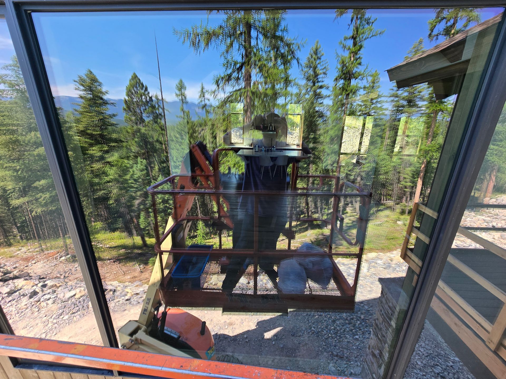 An aerial lift platform with two bags of construction materials sits outside a building window against a forest backdrop.