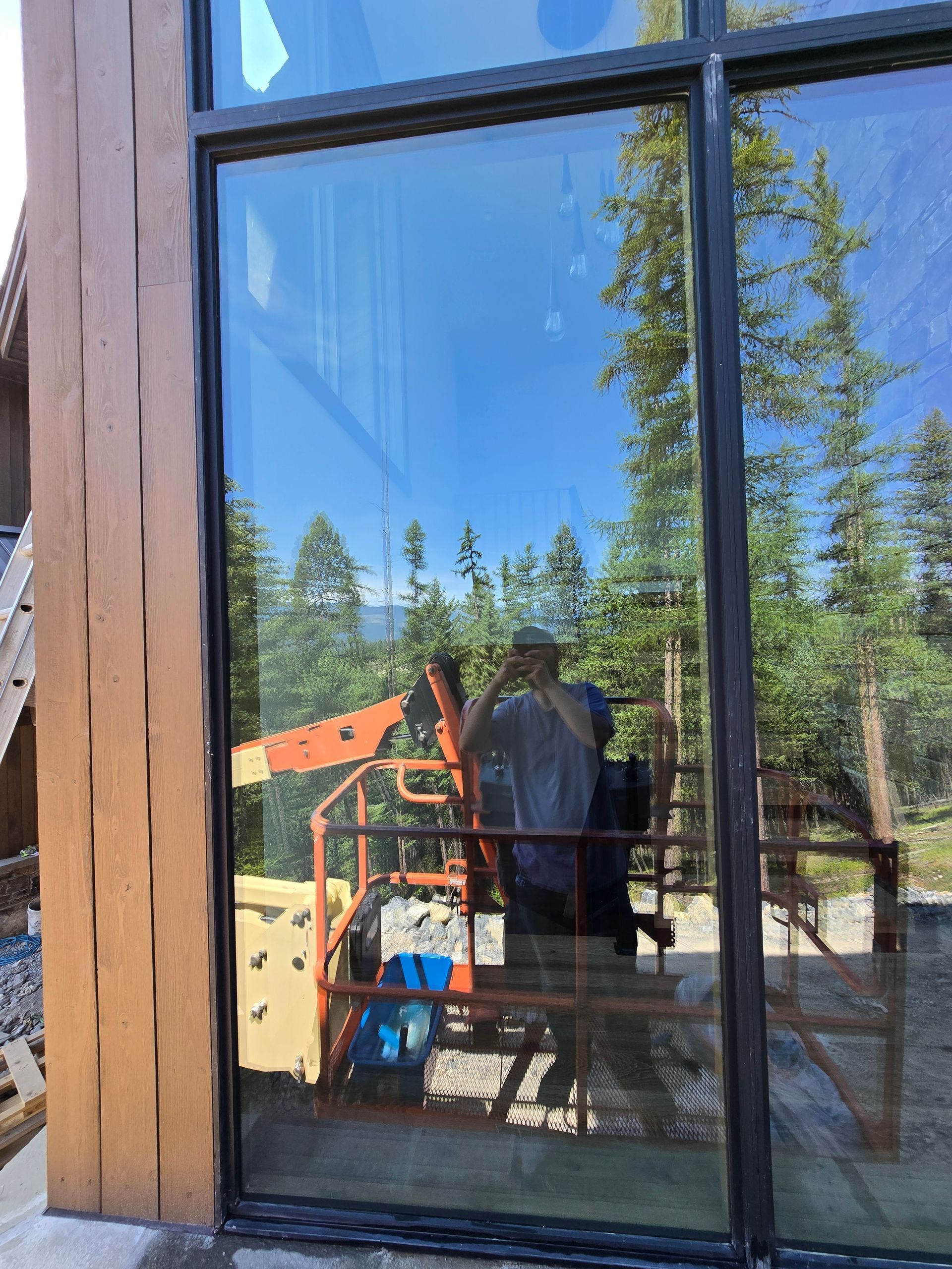 Reflection in a large window showing a person taking a photo in front of construction equipment and pine trees.