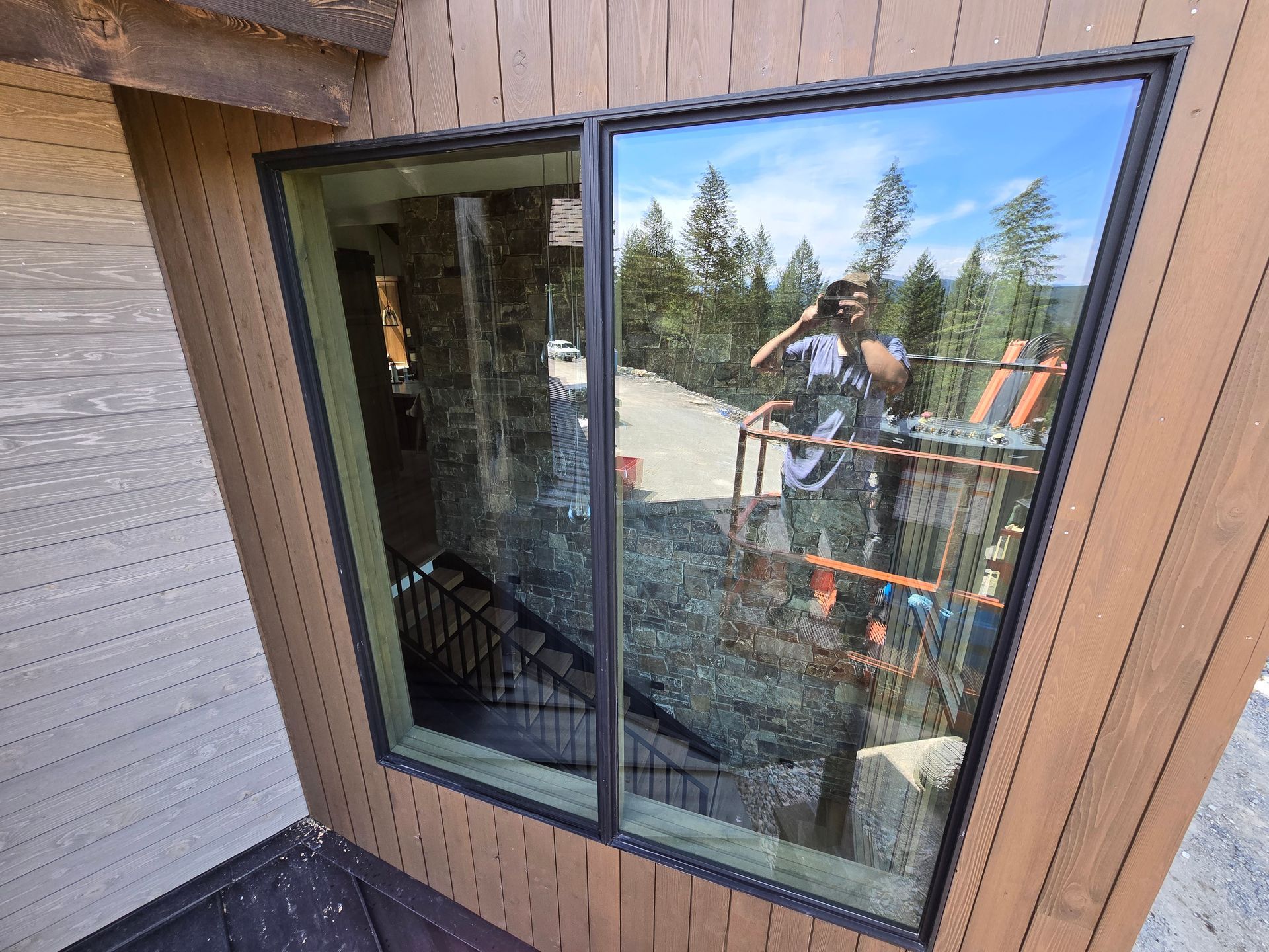 Large, dark-framed window on a wooden exterior wall reflecting a person taking a photo and a construction scaffold.