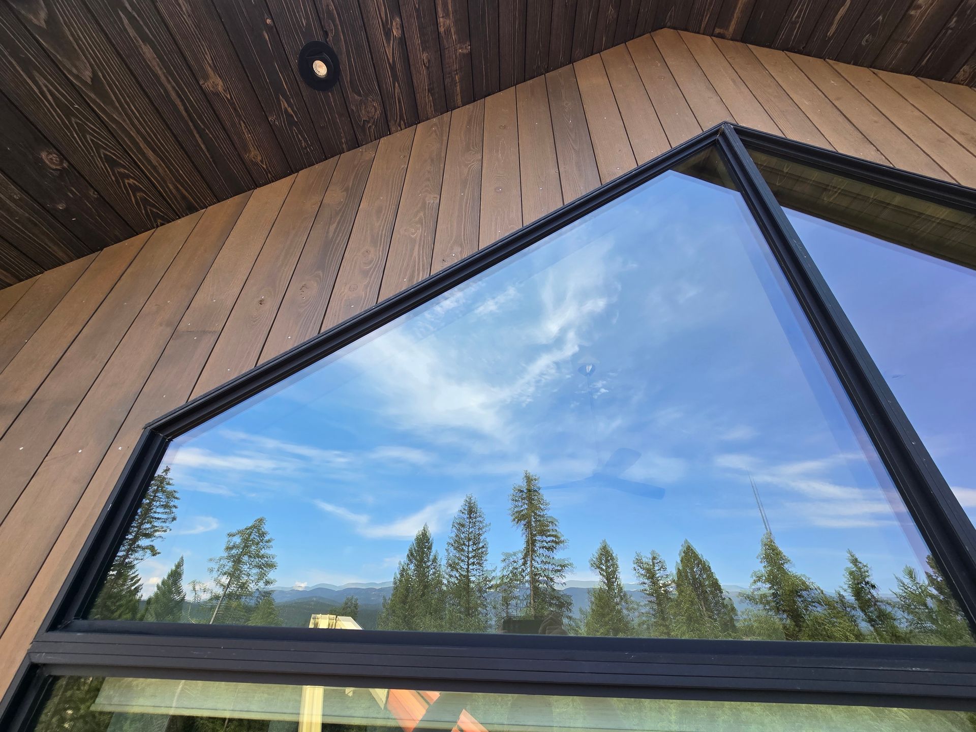 A view through an angled, black-framed window showing a forest and mountain landscape under a bright, cloudy blue sky.