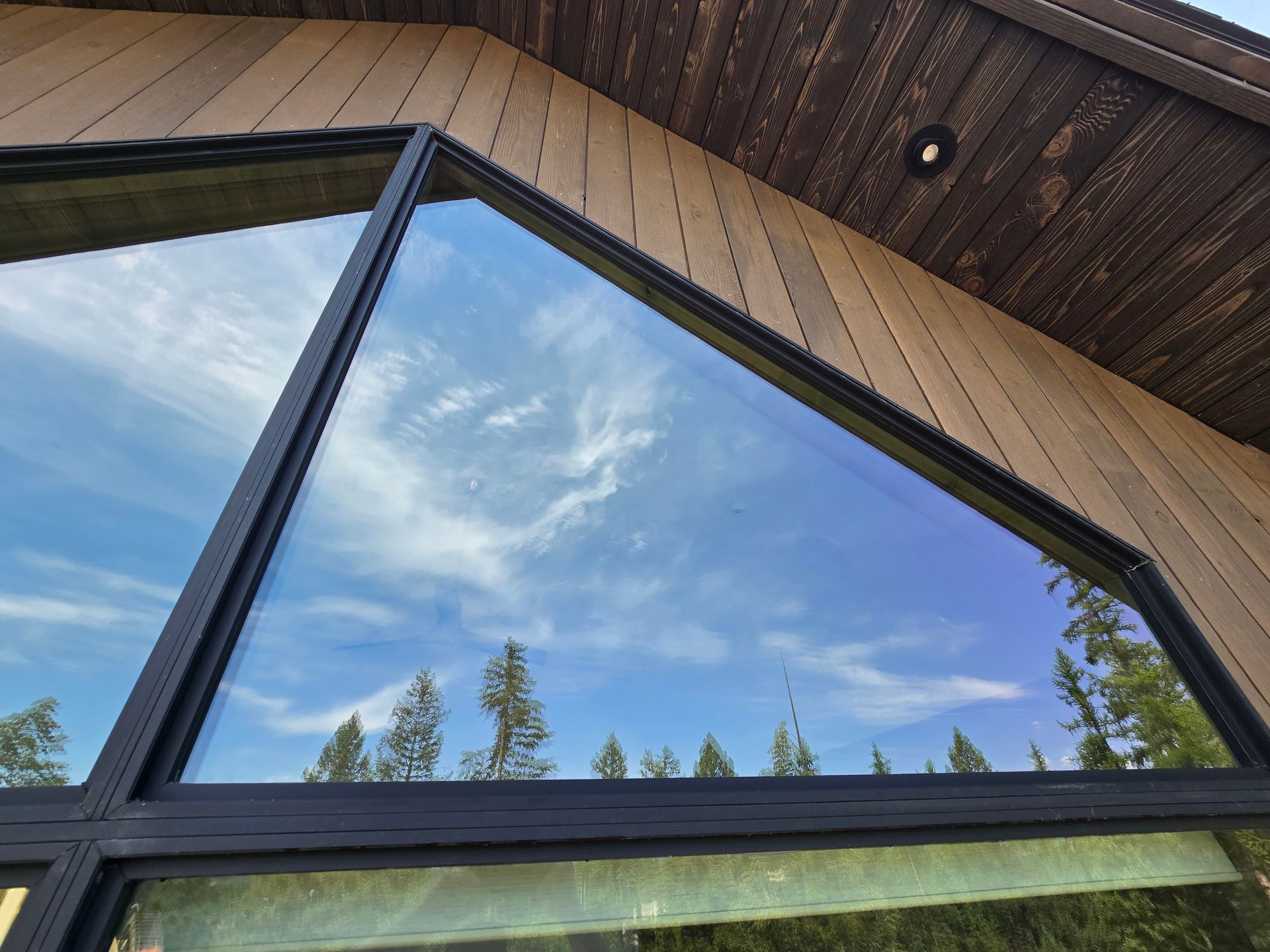 An angular, black-framed window on a modern building reflects a blue, cloudy sky and surrounding pine trees.