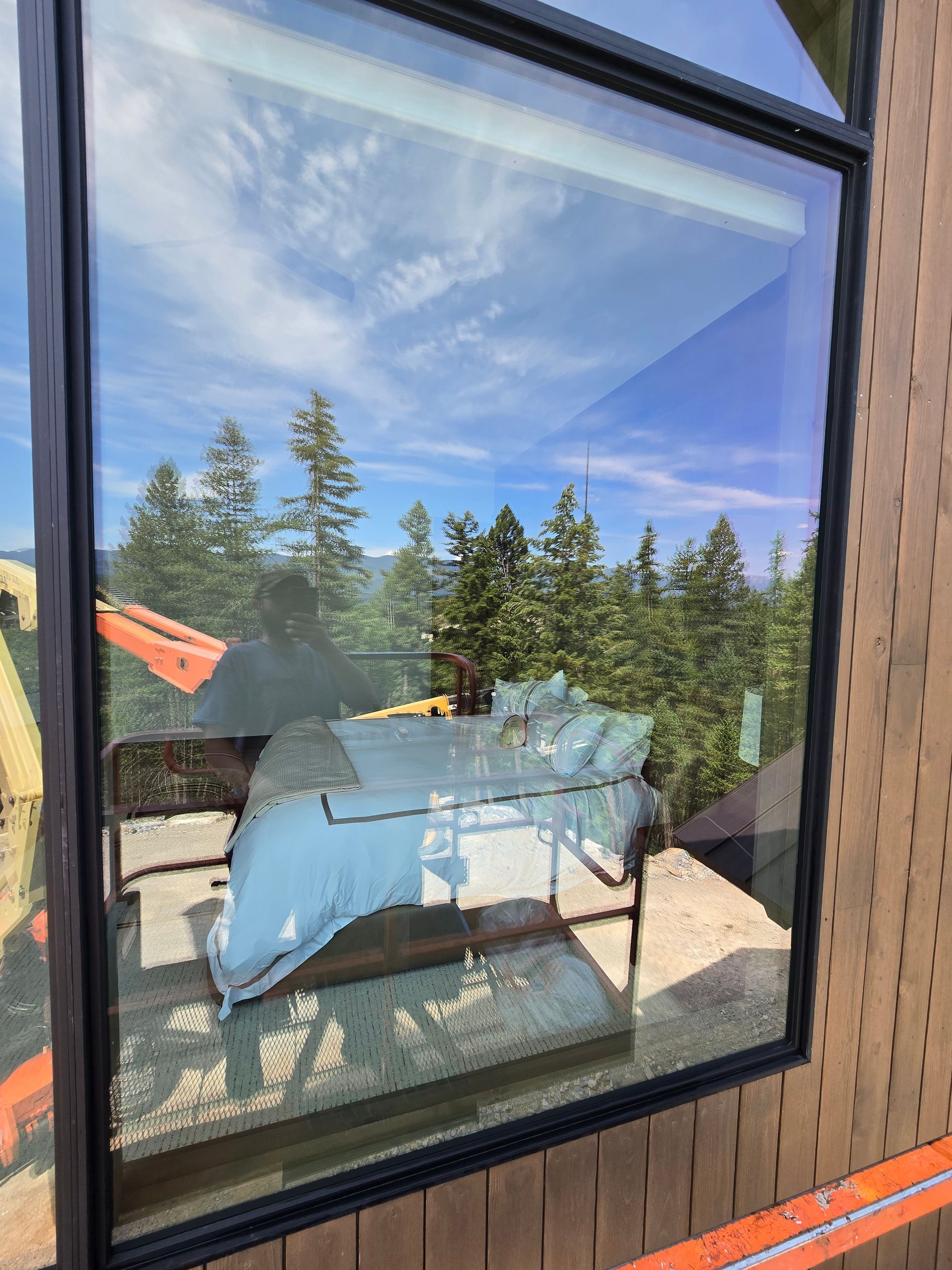 A window with a reflection showing a white bed and forest landscape, with orange construction framing visible outside.