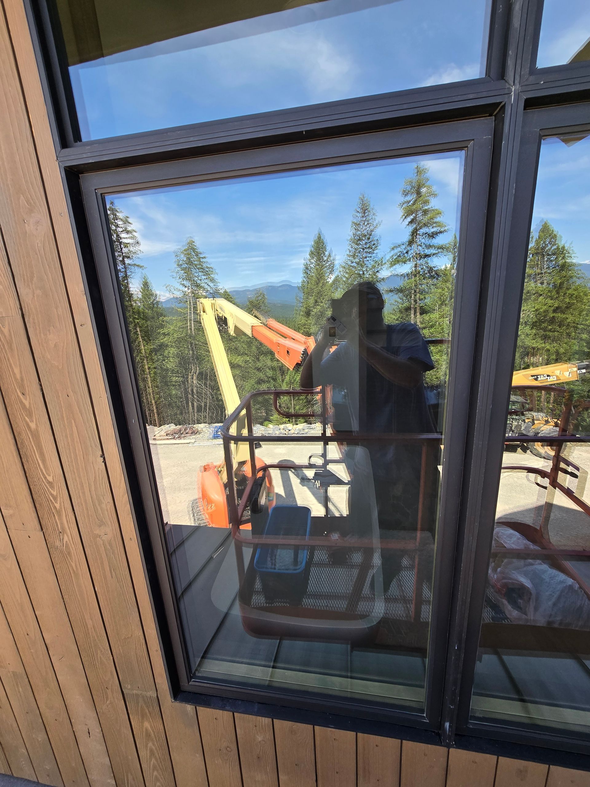 Reflected in a window are a person in a construction lift, an orange boom arm, trees, and a mountain landscape.