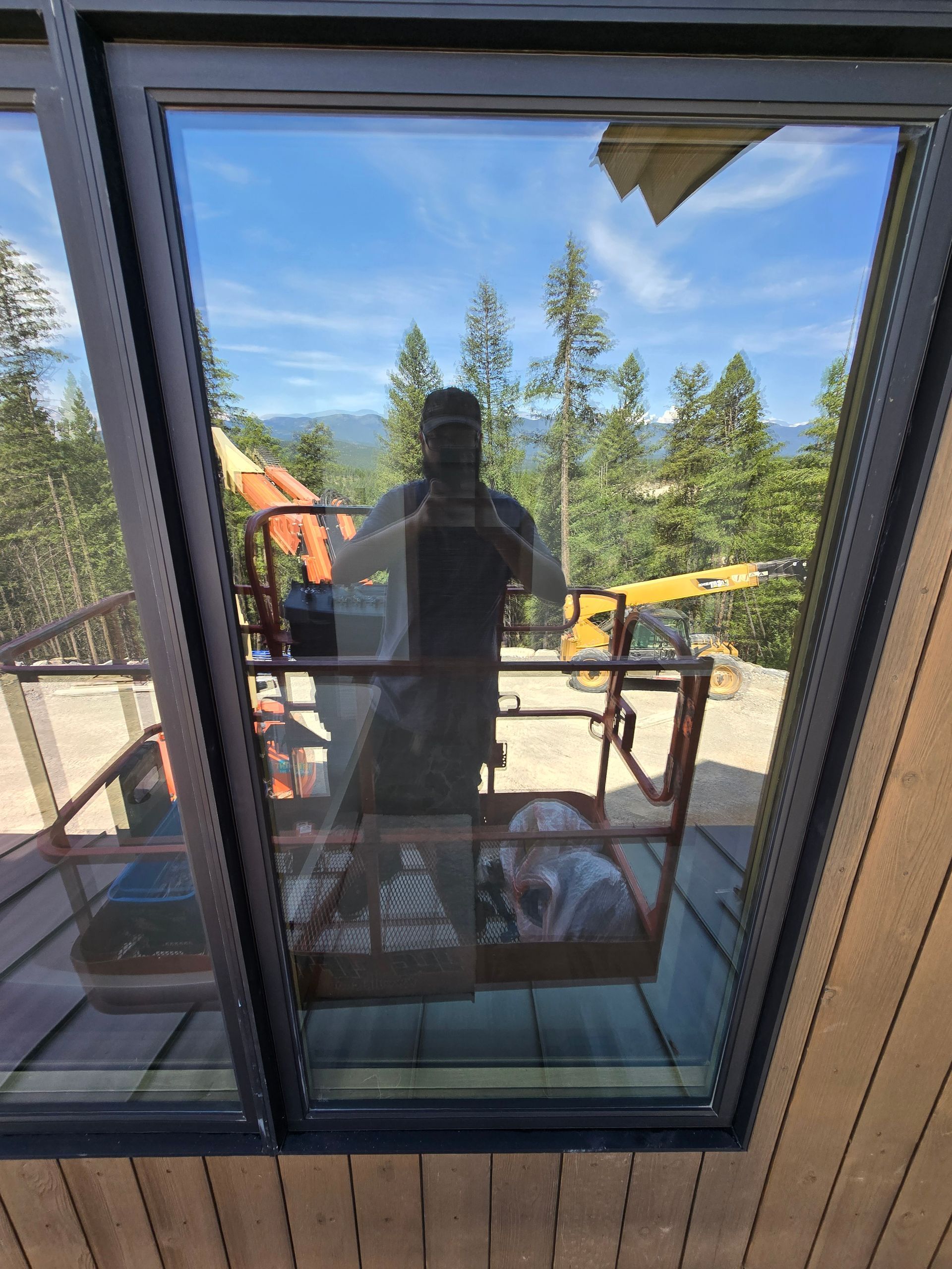 A person reflected in a window showing a construction site with a yellow excavator against a backdrop of trees.