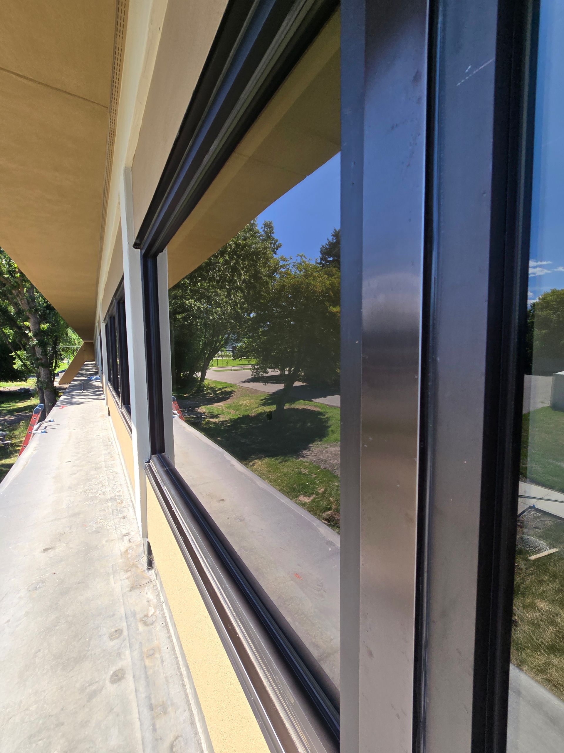A view from an outdoor walkway looking along a building wall with large glass windows reflecting trees and a clear blue sky.