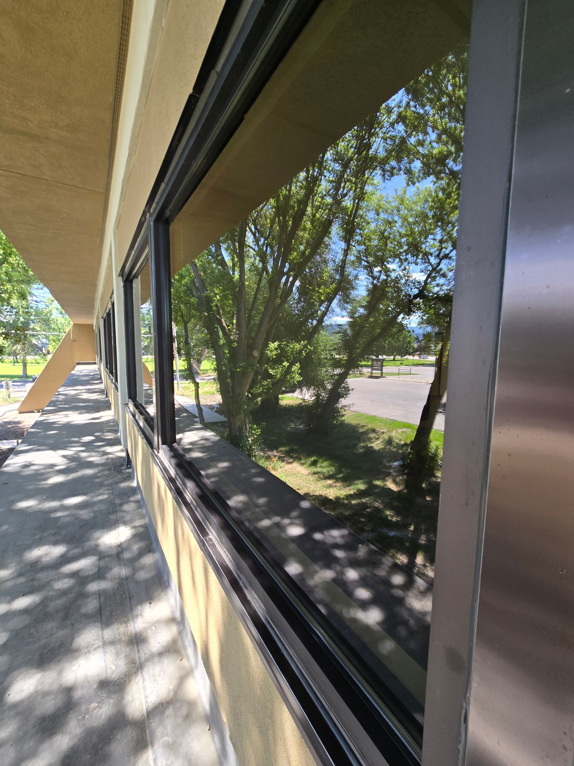 A sidewalk runs along a tan building with large, dark windows reflecting nearby trees and a parking lot.