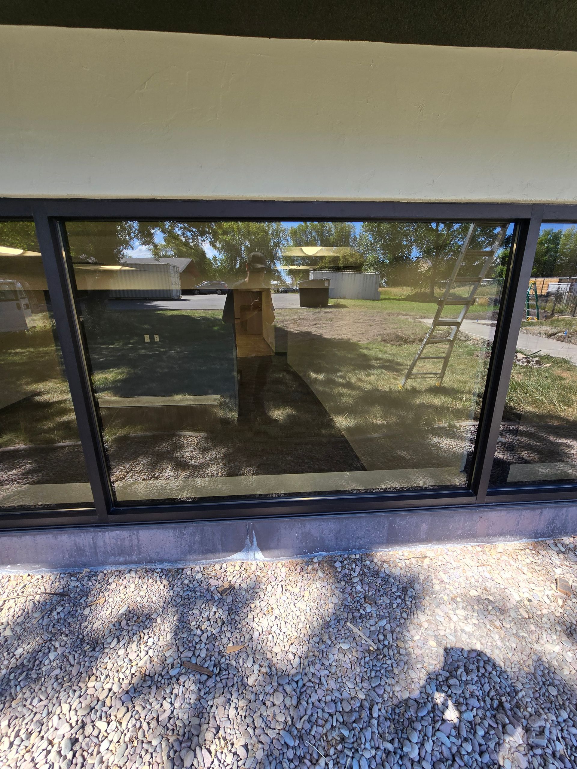 A large, dark-framed window on a building exterior, reflecting the surrounding trees and ground covered in light gravel.