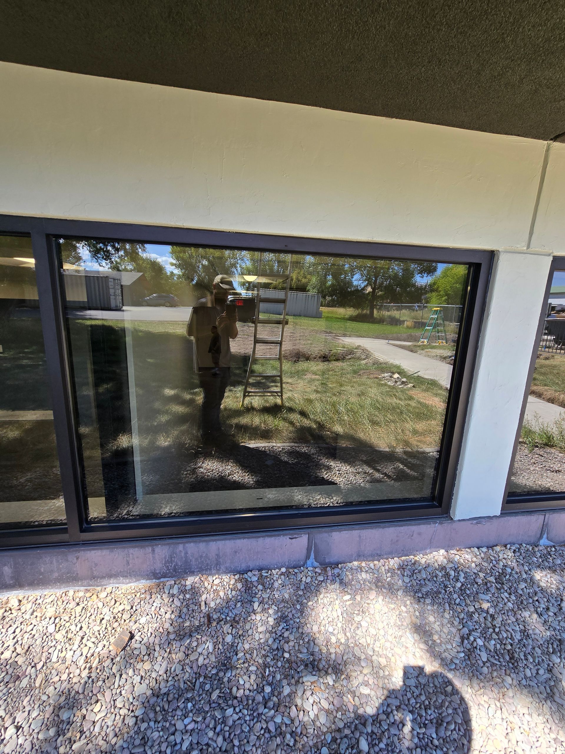 A dark-framed window reflects a ladder standing on gravel, with trees and a sidewalk visible in the background.
