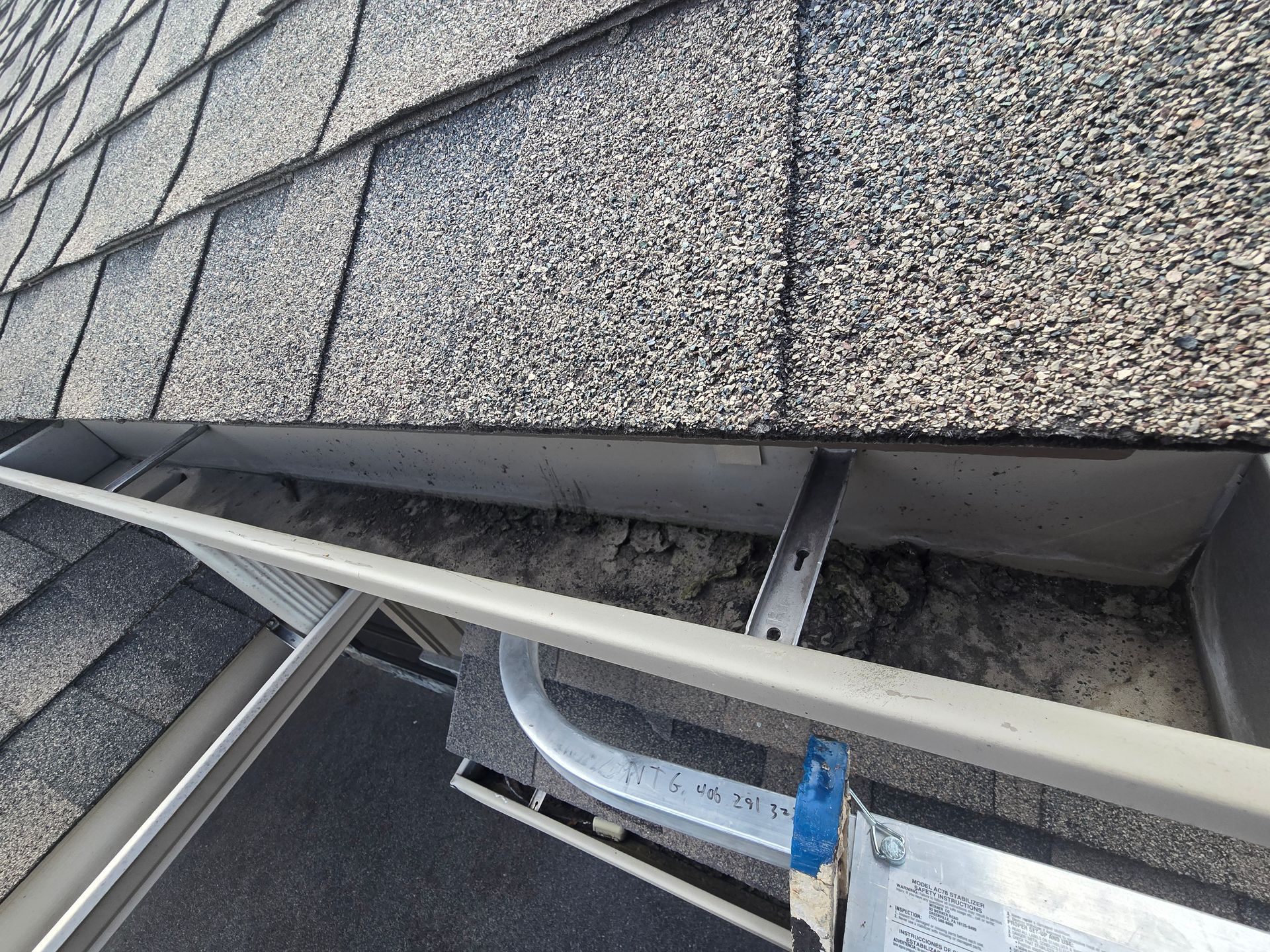 An elevated view of a gutter filled with dark debris, showing the roof shingles above and a metal ladder in the foreground.
