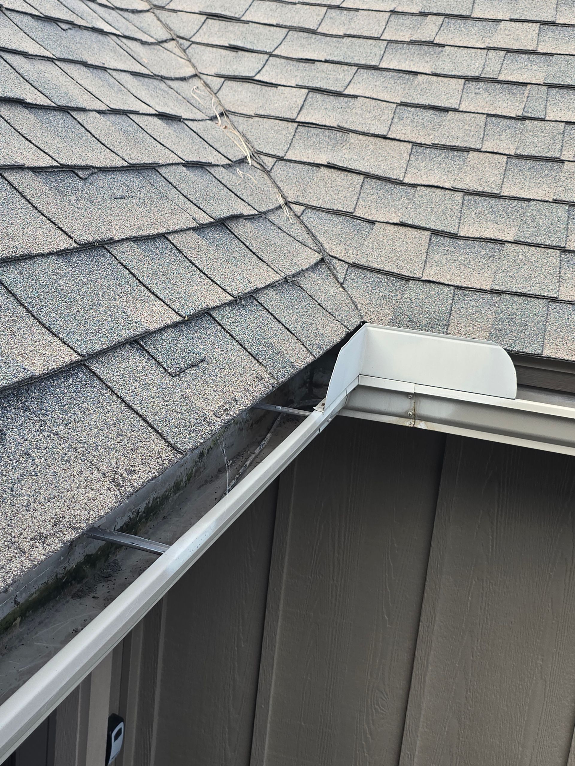 A white metal gutter corner piece installed at the intersection of two roof sections with grey asphalt shingles.