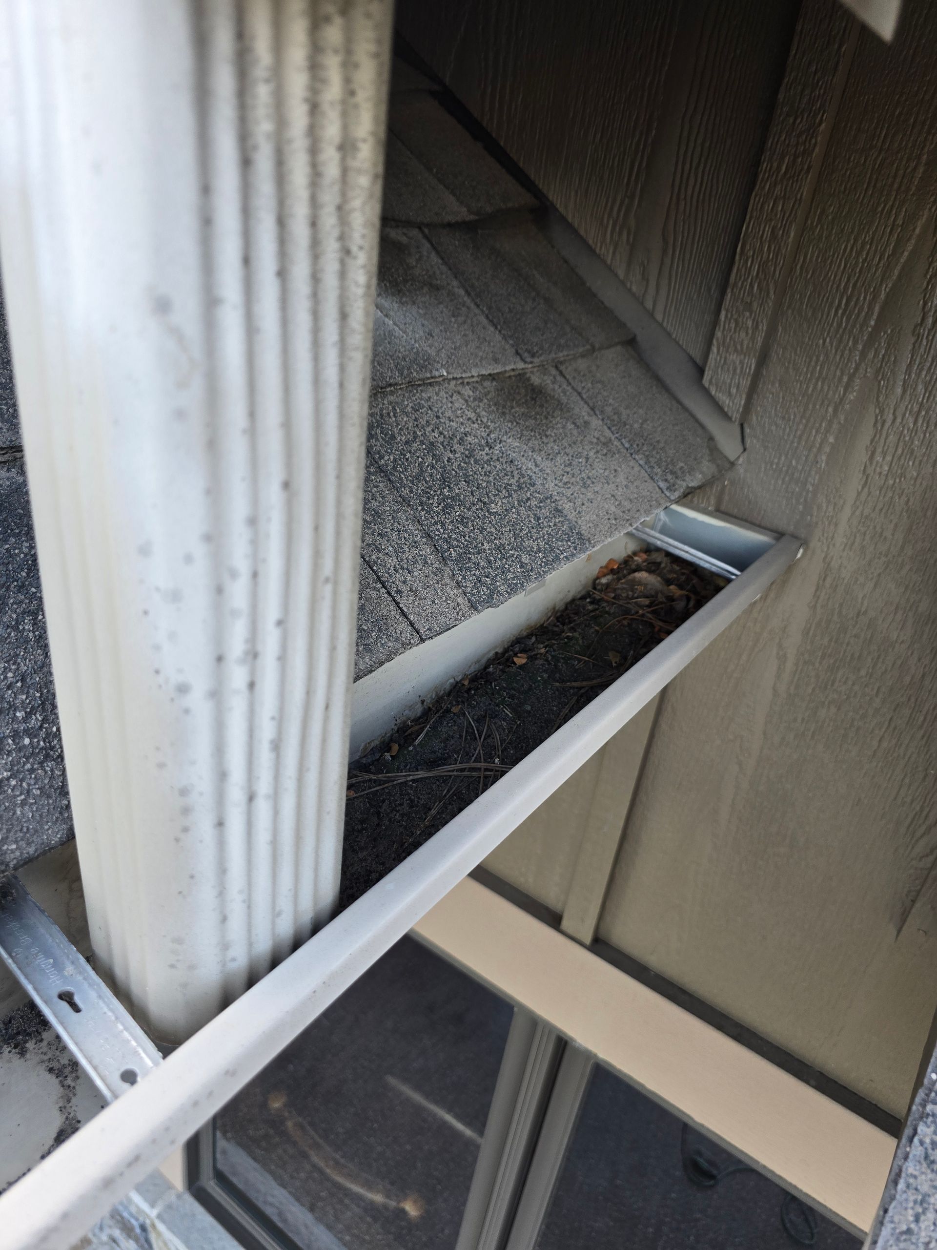 A white gutter downspout next to a gutter section filled with dark debris and shingle grit against a siding wall.