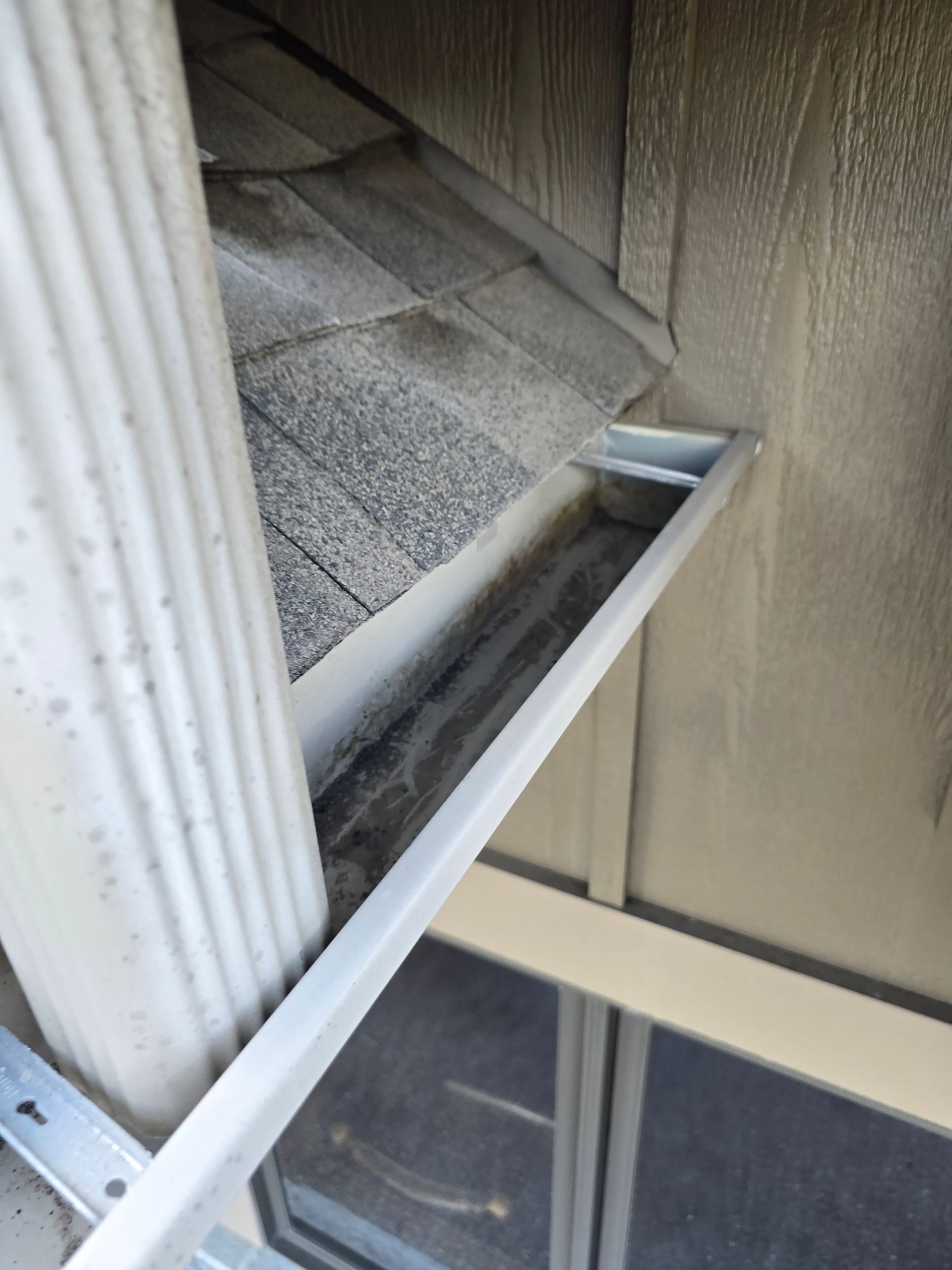 A close-up view of a metal roof gutter attached to beige siding, with a vertical downspout visible on the left side.