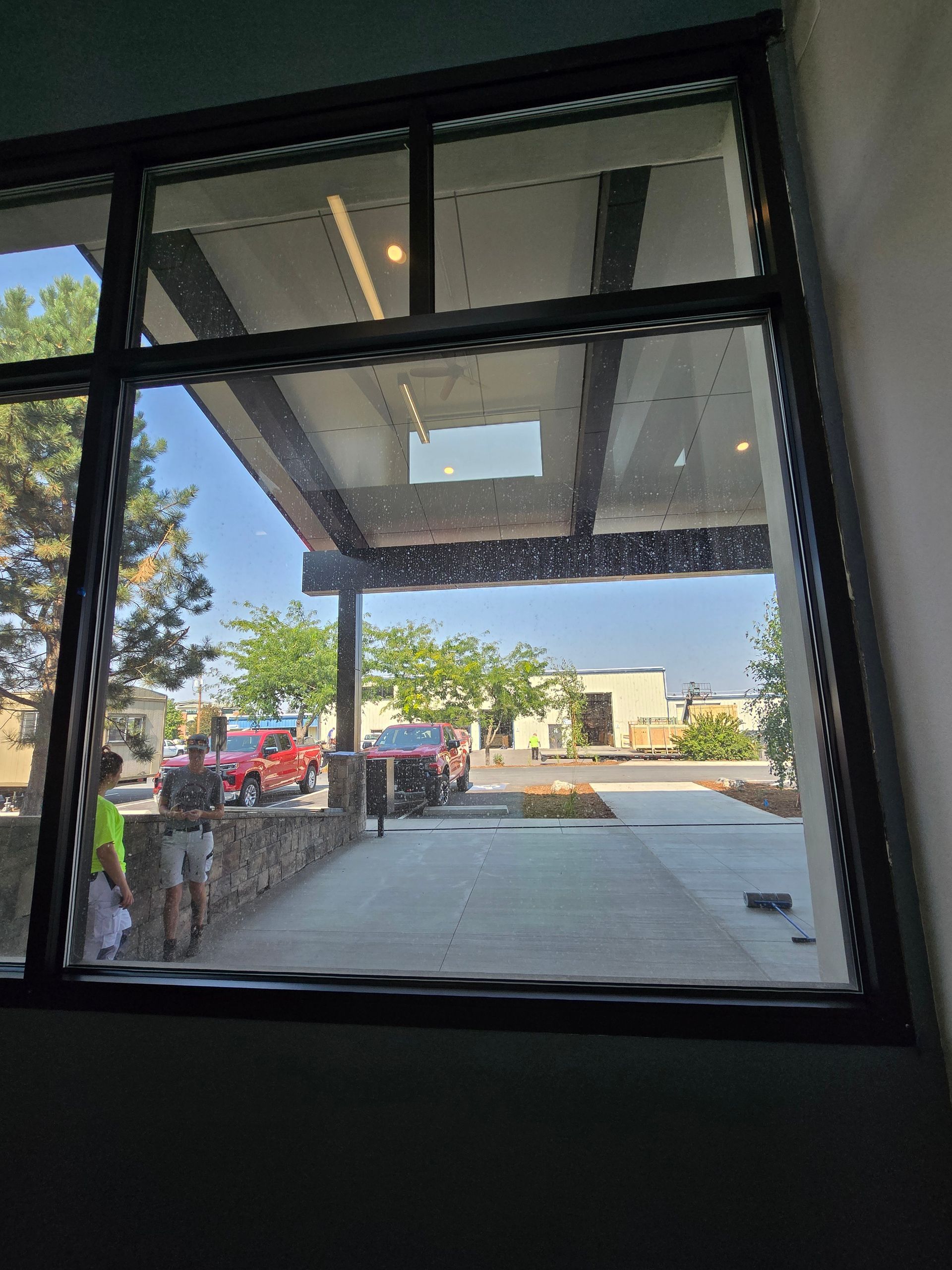 View from inside through a large window showing a parking lot, trees, and people under an outdoor overhang on a sunny day.