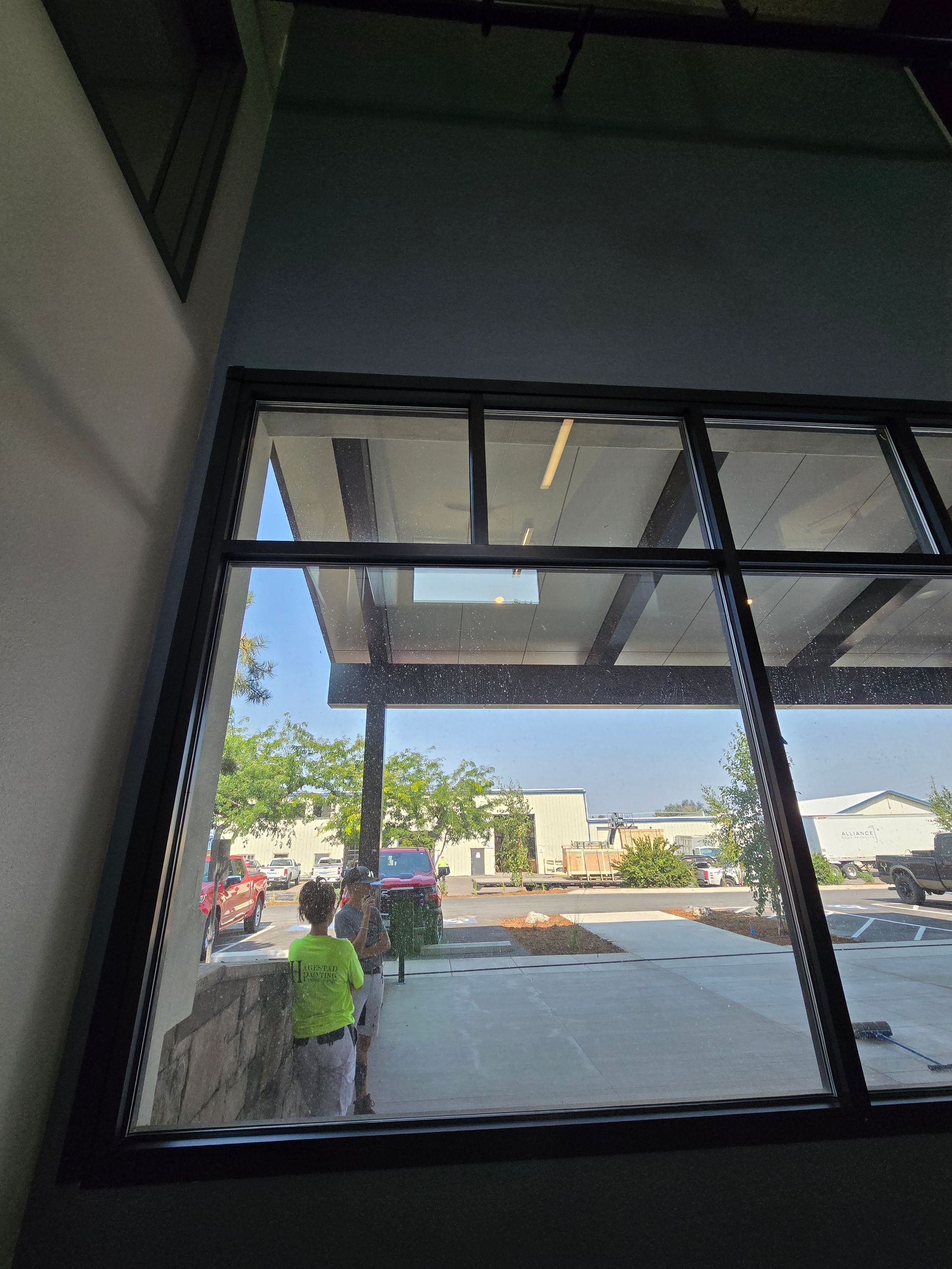 A child in a bright yellow shirt stands outside a large, multi-paned window looking toward a parking lot on a sunny day.