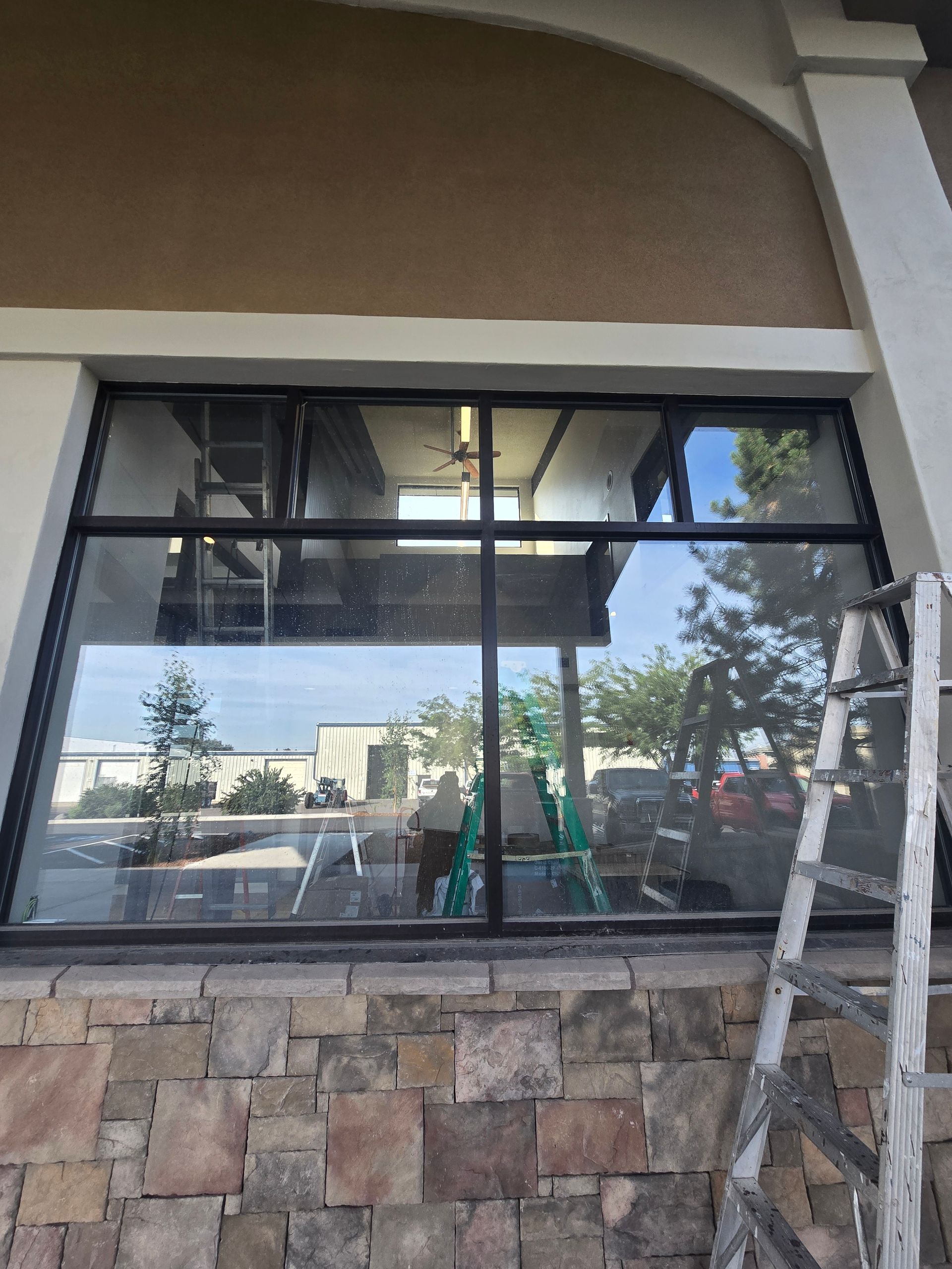 A commercial storefront window with black frames, reflecting the parking lot, with a ladder leaning against the right side.