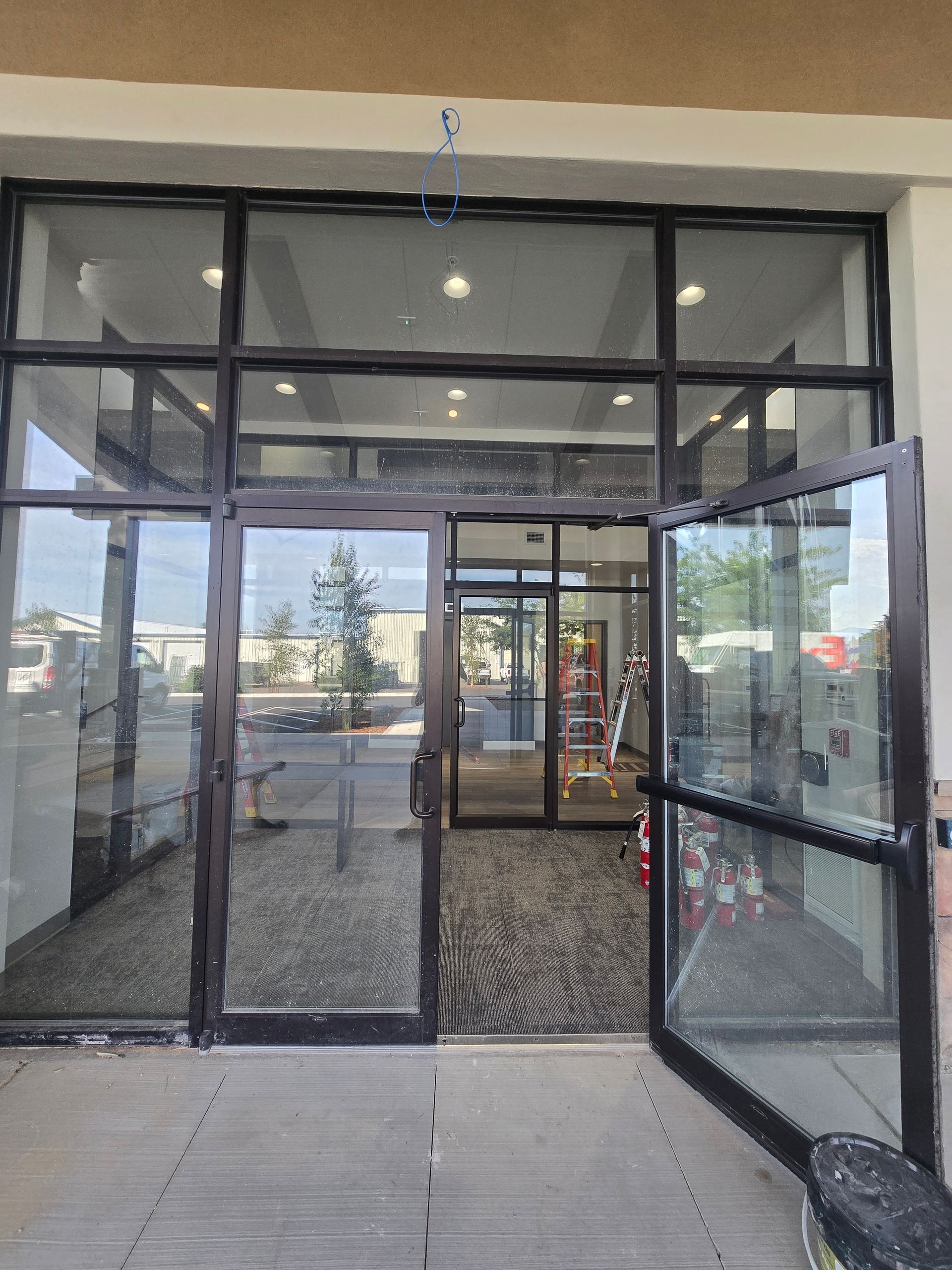 A glass entrance with double doors open to a store interior featuring gray carpet, recessed lighting, and a ladder.
