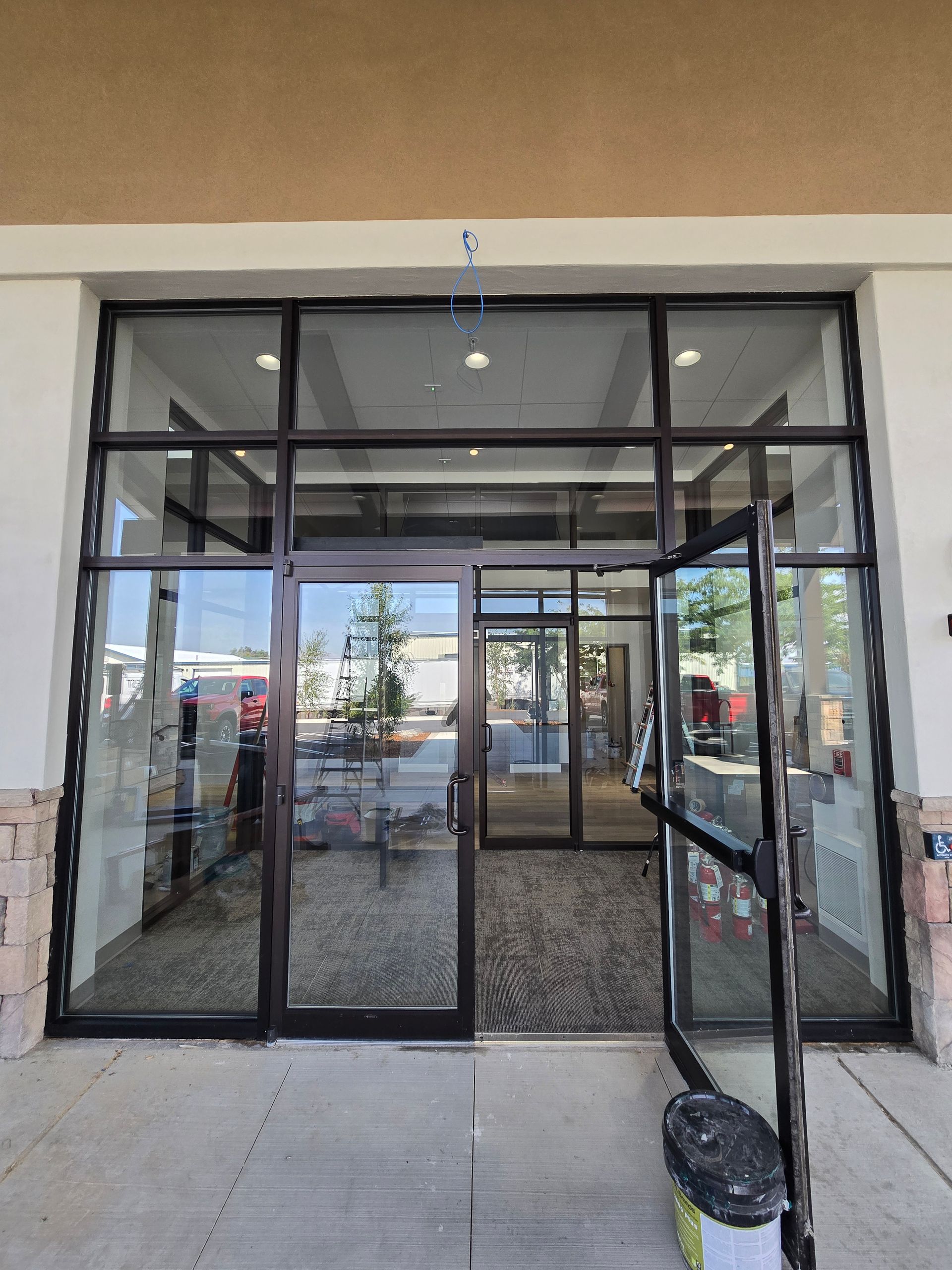 Entrance to a storefront with glass doors and windows, featuring a dark metal frame and a bucket on the sidewalk.