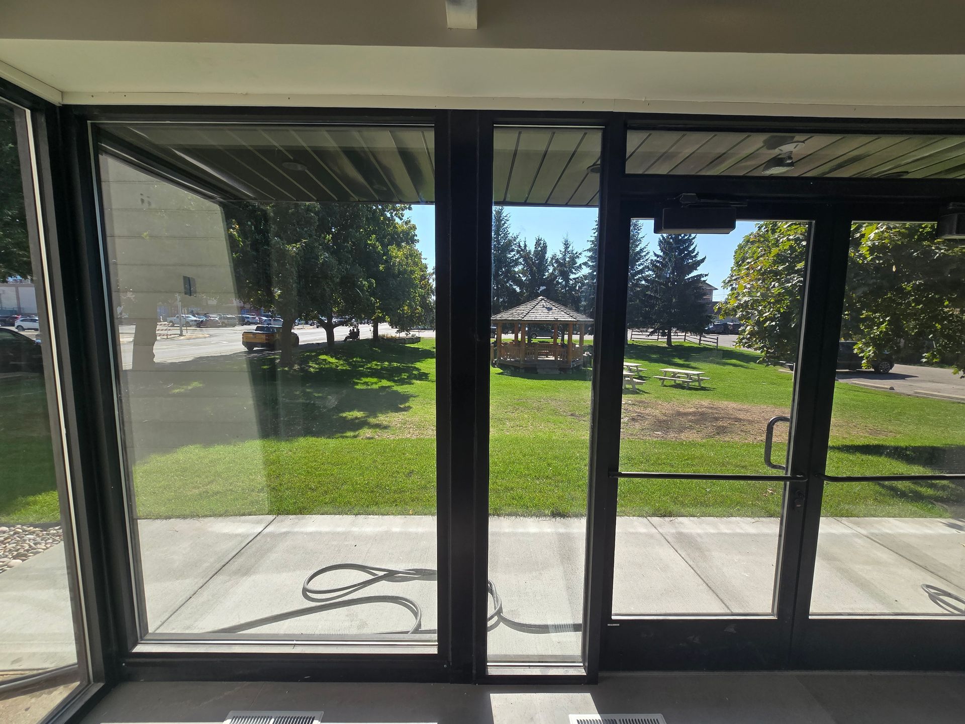 A view through glass doors at an entrance looking out onto a sunny lawn with a wooden gazebo and scattered trees.