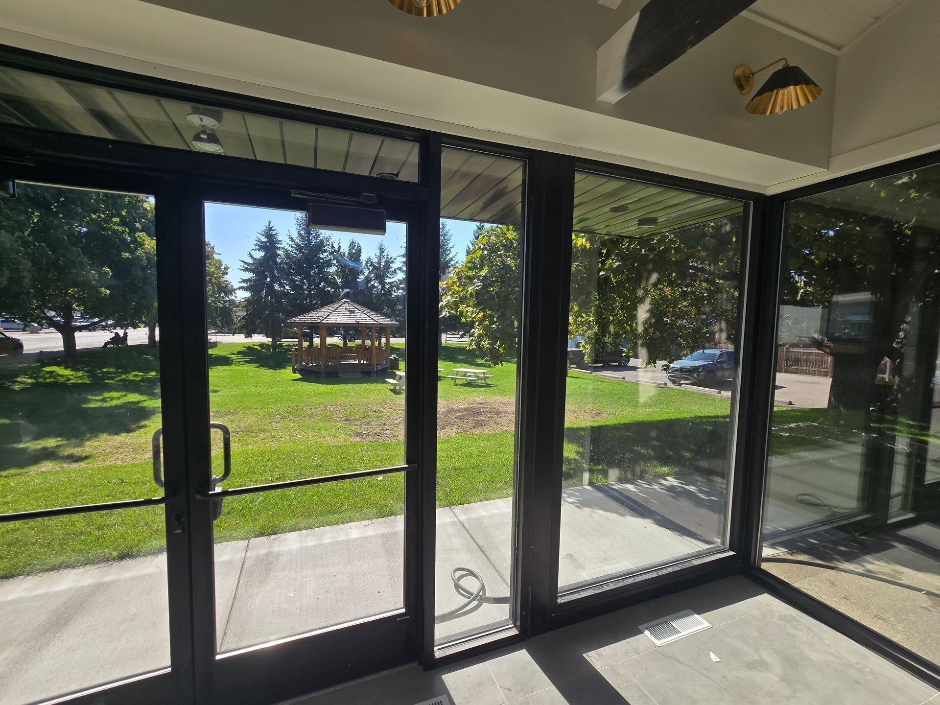 A view through a glass door and window looking out onto a grassy park with a wooden gazebo under a clear blue sky.