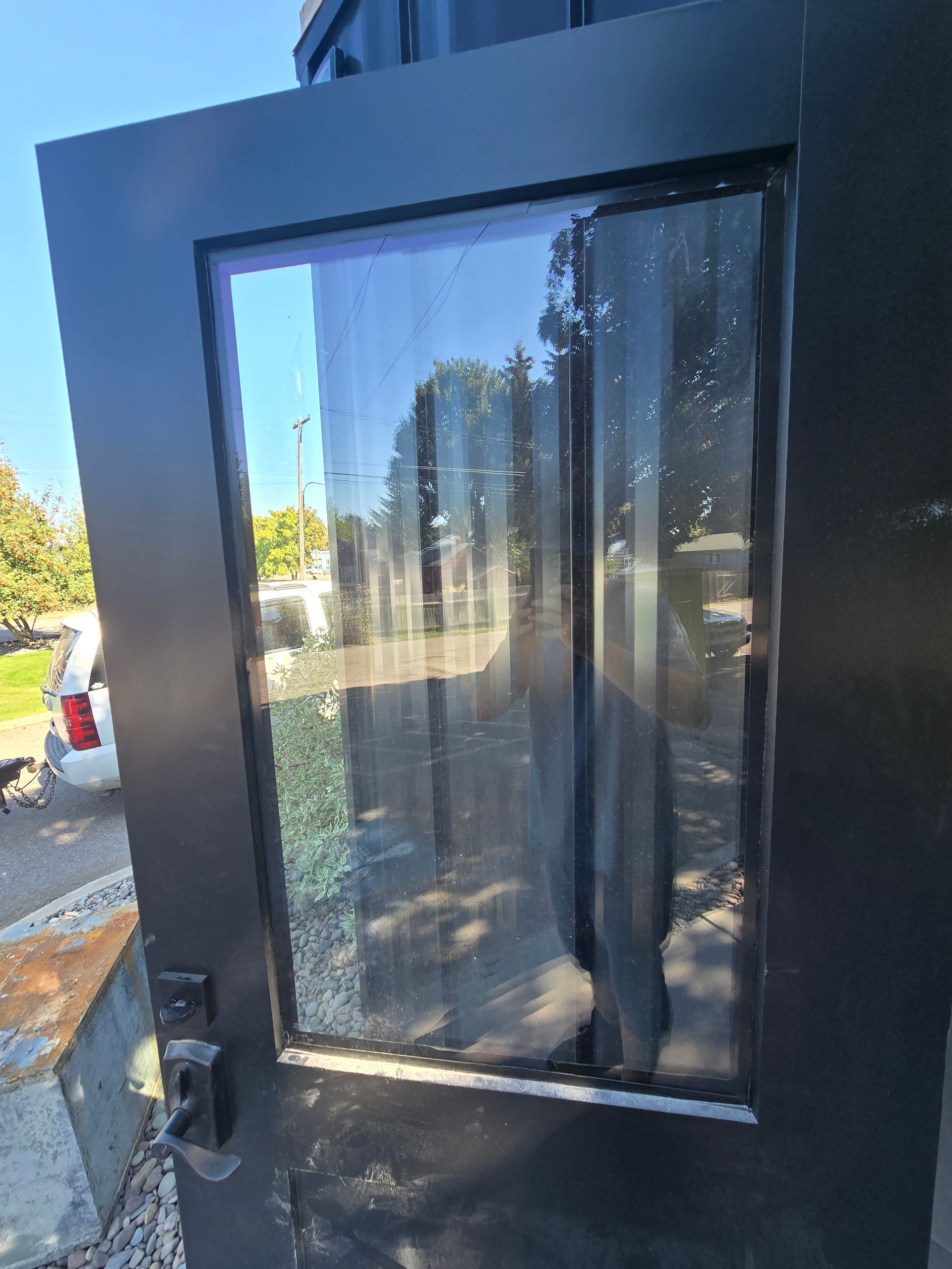 Black metal door with a large glass window pane, reflecting trees and a white vehicle in the background.