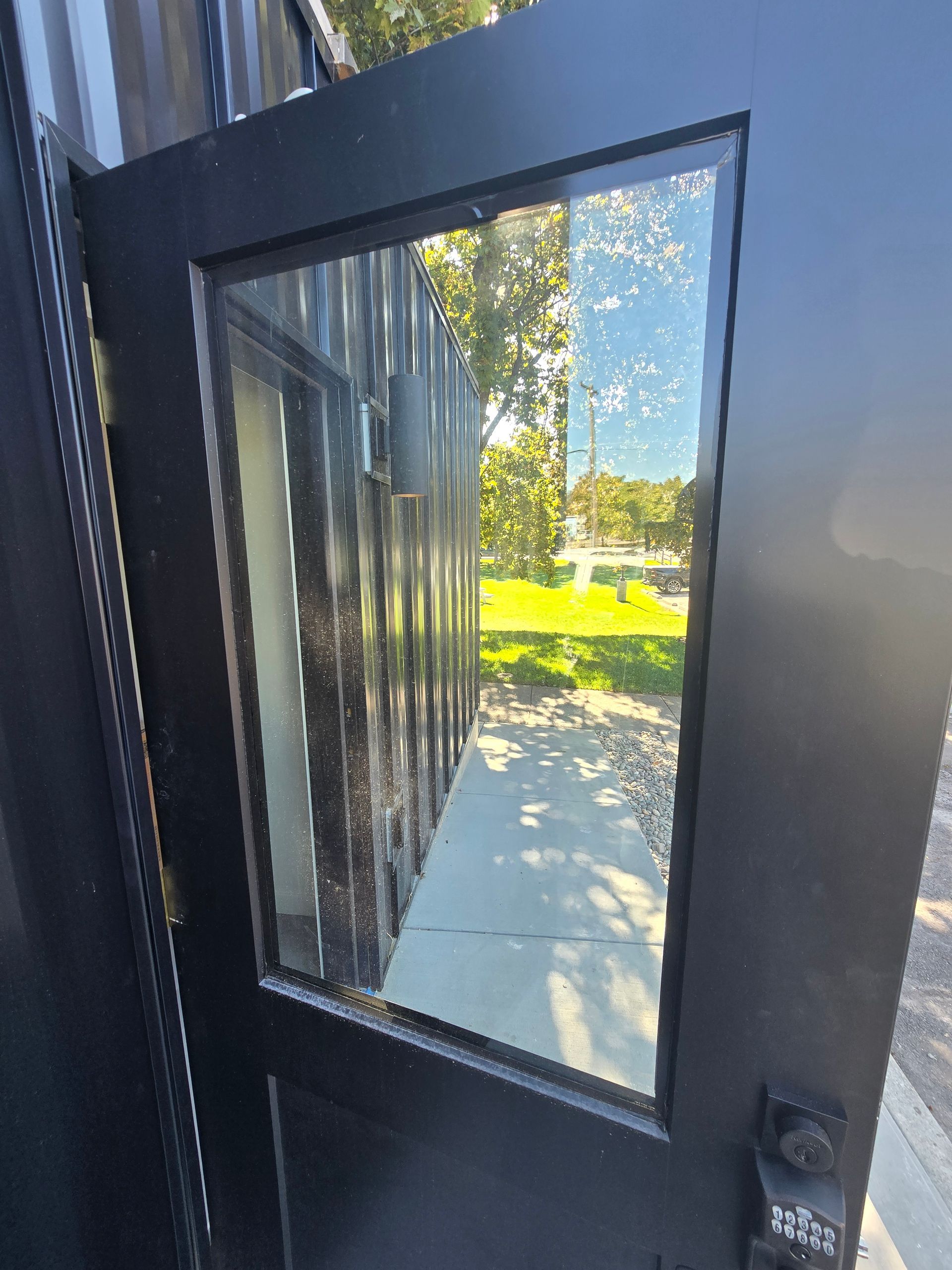 A black door with a large glass pane, reflecting a building exterior, trees, and grass.