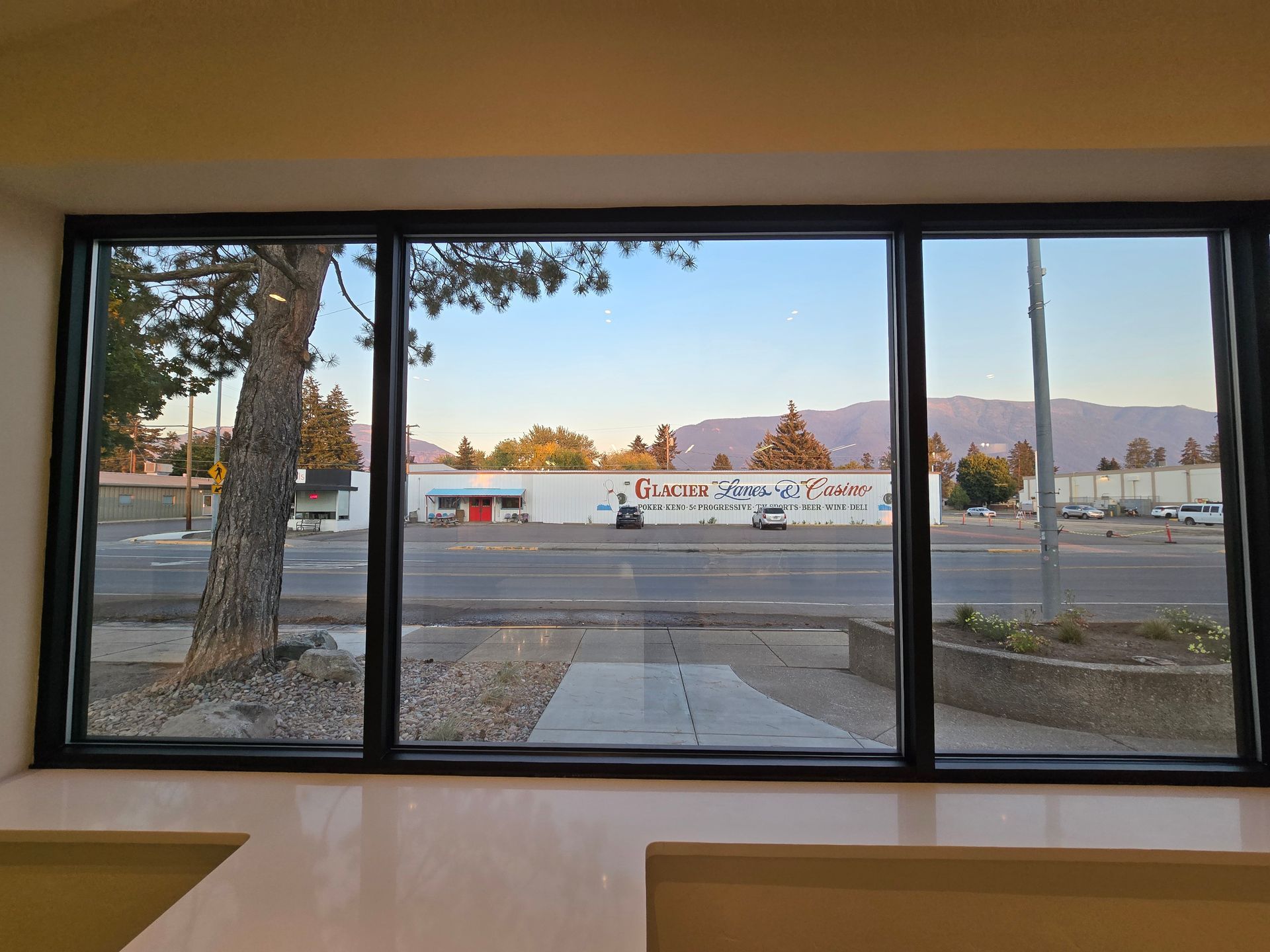 A view from inside through a black-framed window looking out at a street, a parking lot, a commercial building, and hills.