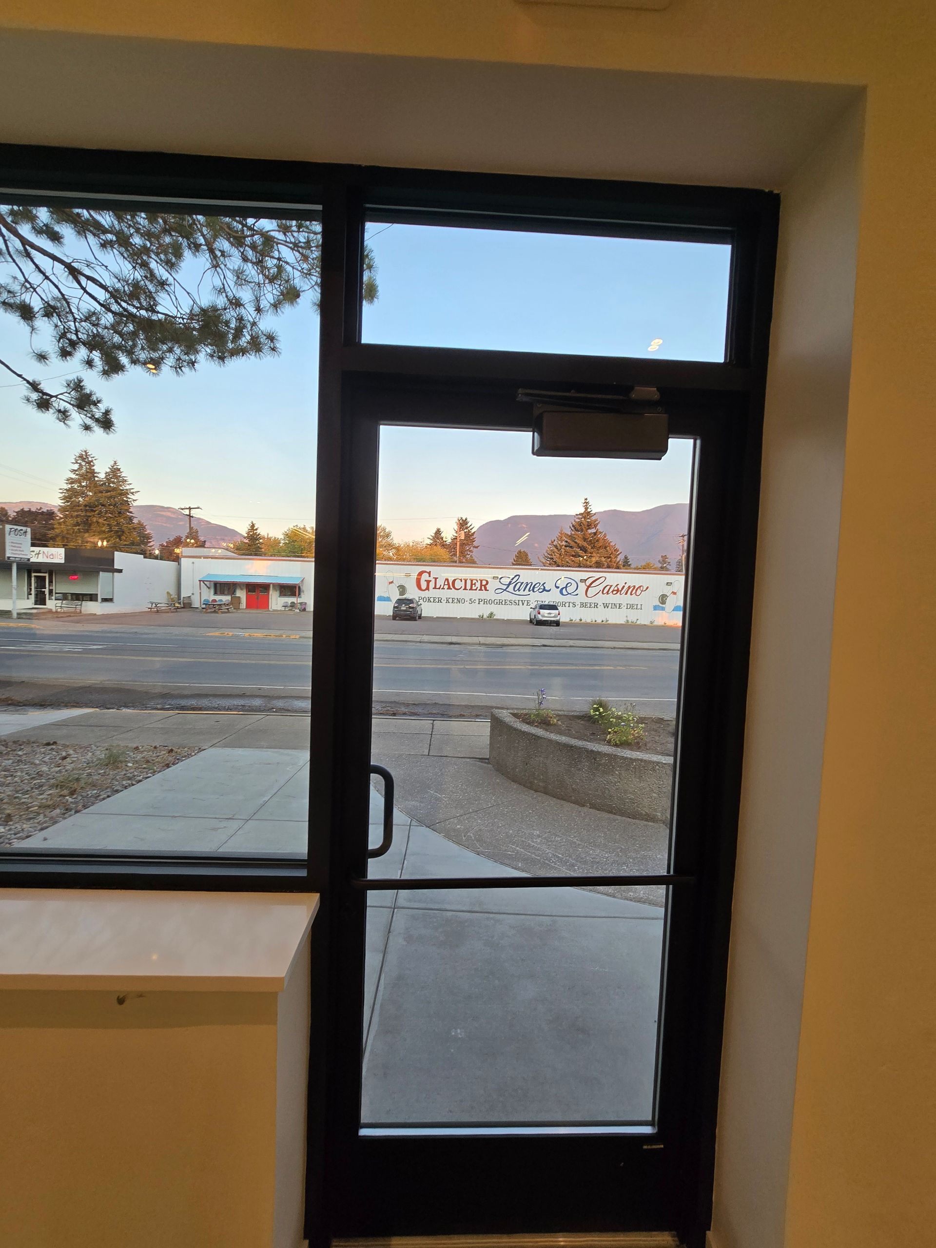 An interior view looking out through a black-framed glass door and side window at a parking lot, mural, and mountains.