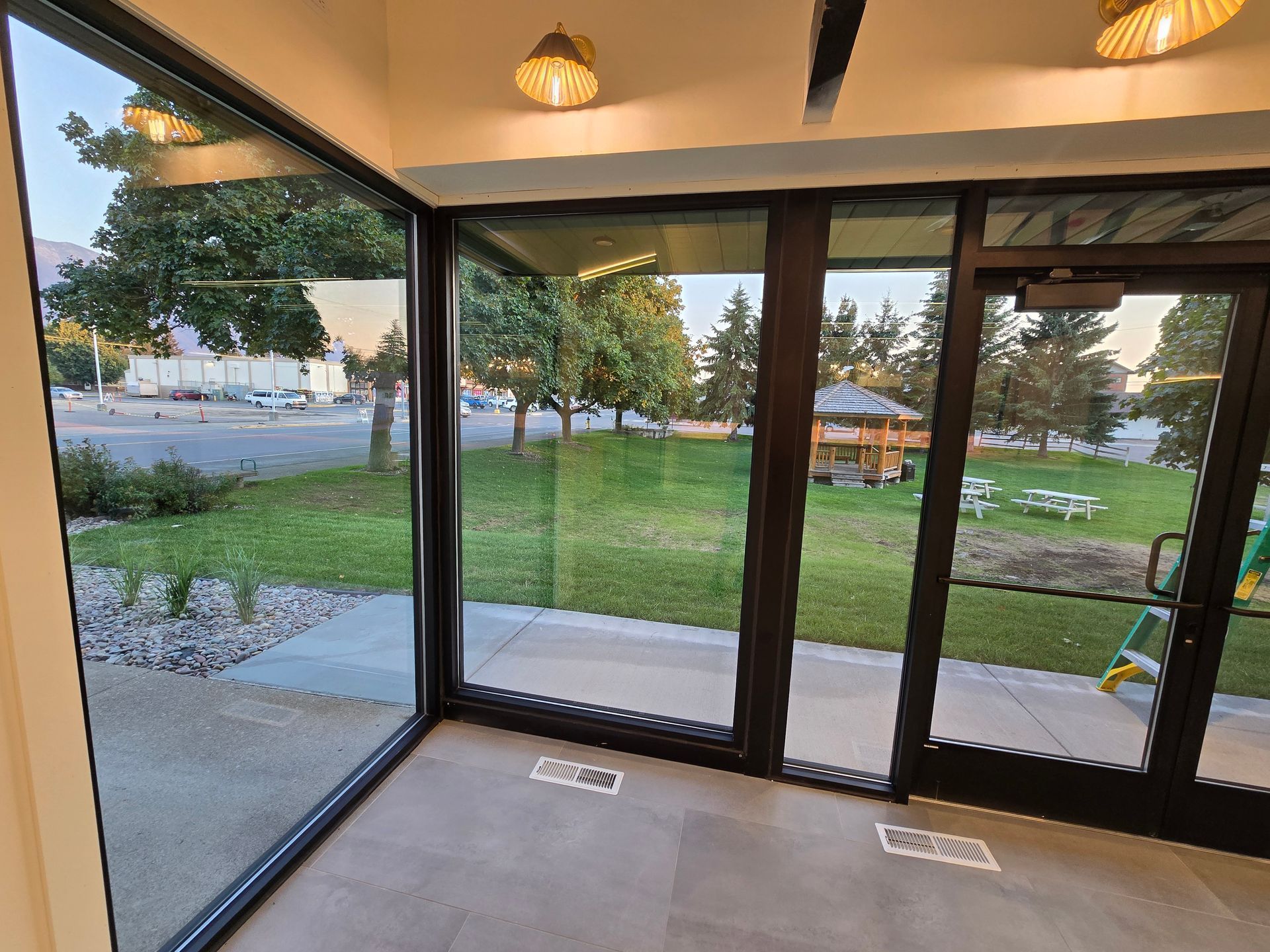 A view through floor-to-ceiling windows looking out onto a grassy courtyard with trees, a small wooden structure, and sky.