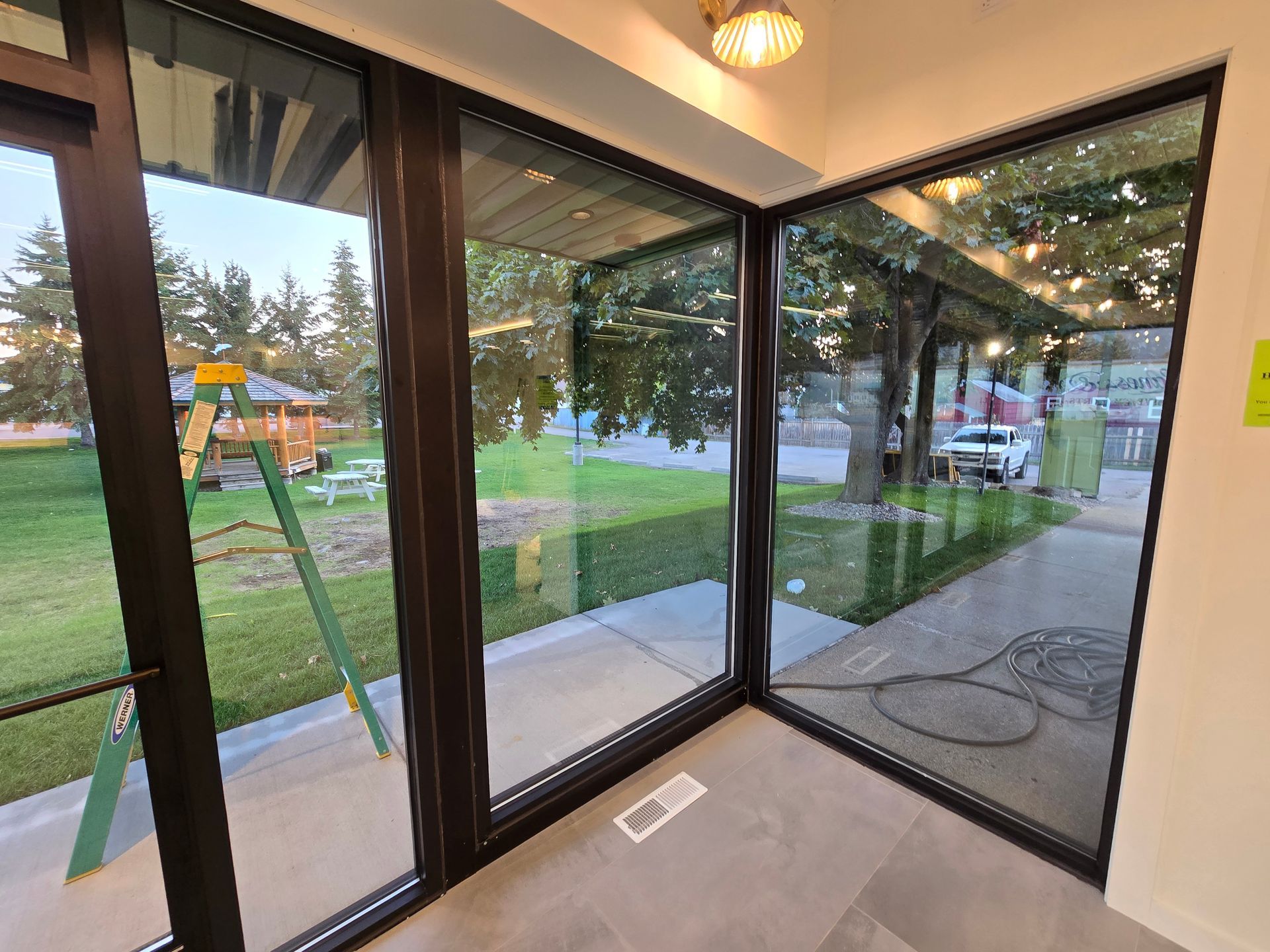 View from indoors through large glass corner windows looking out at a grassy lawn, trees, and a distant building.