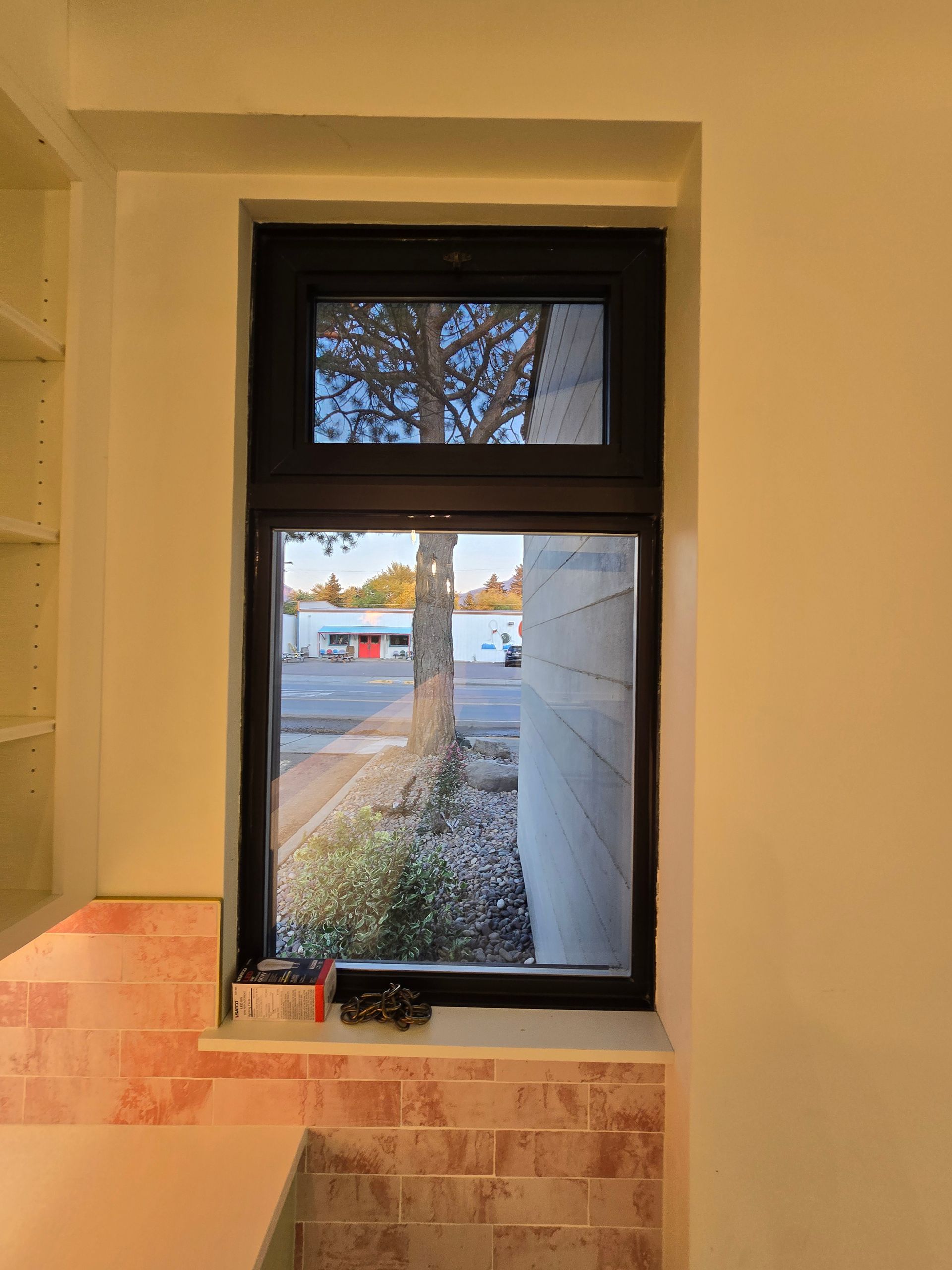 A kitchen window with a dark frame set in a wall with pink subway tile, overlooking a street and trees at sunset.