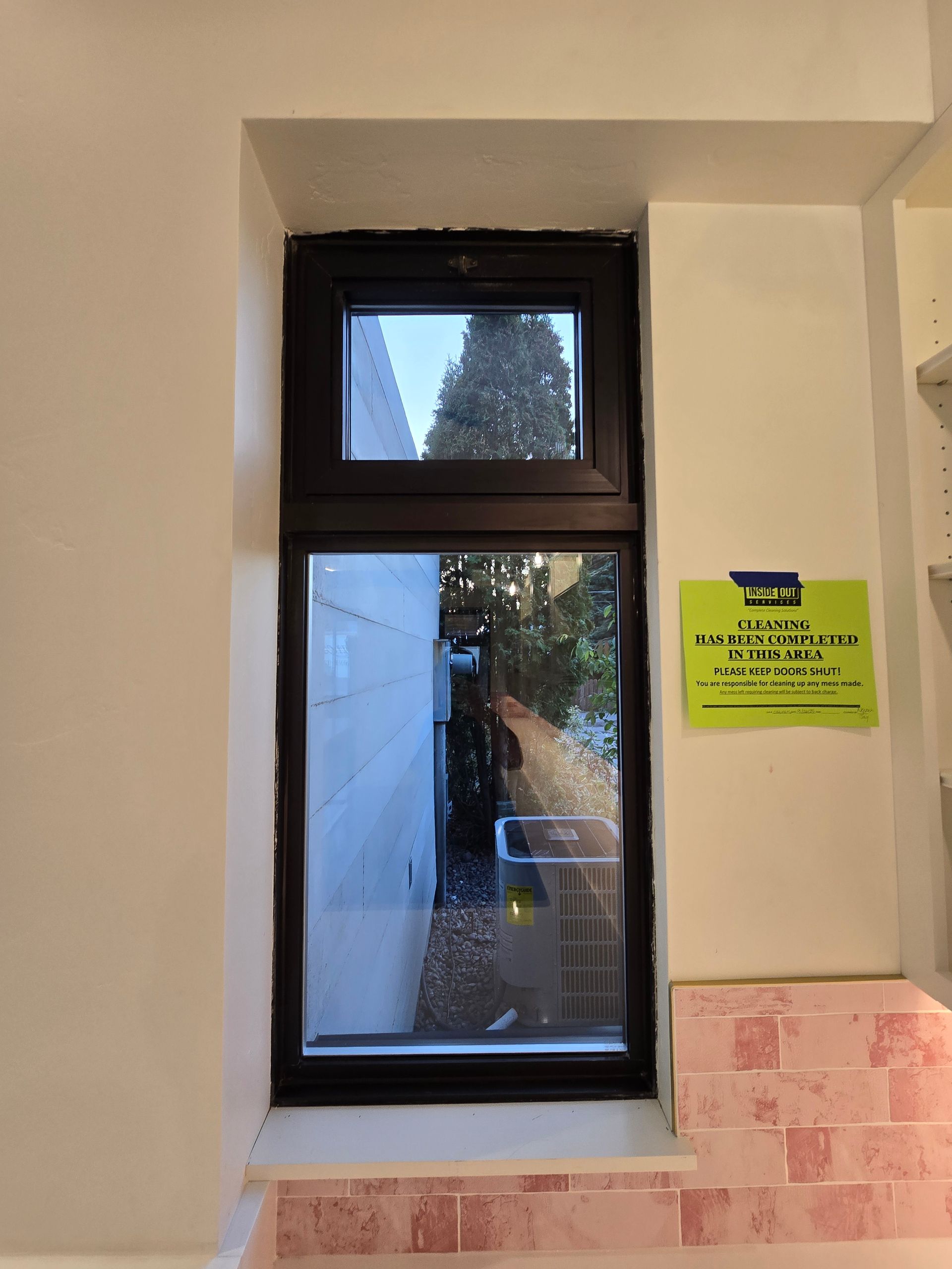 A dark-framed, two-pane window set in a white wall above pink tiles, with a green notice posted on the right side.