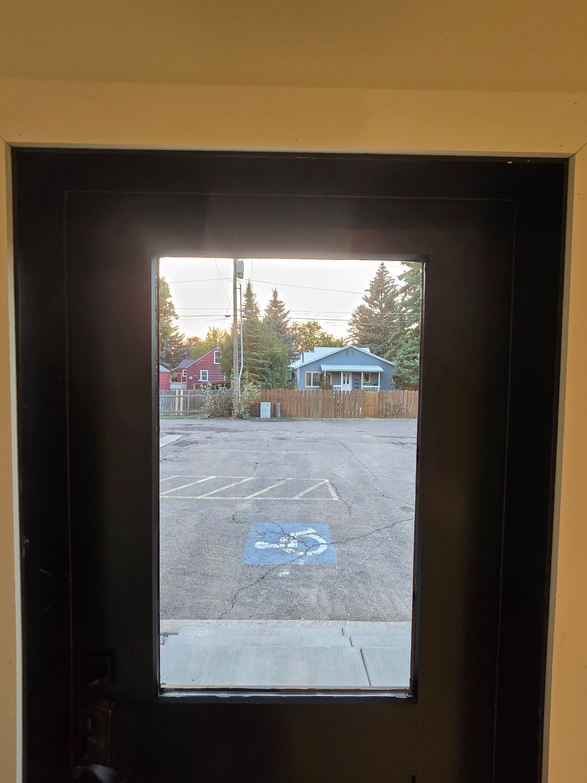A view through a black door window shows a gravel parking lot with a handicap parking symbol and a house in the distance.
