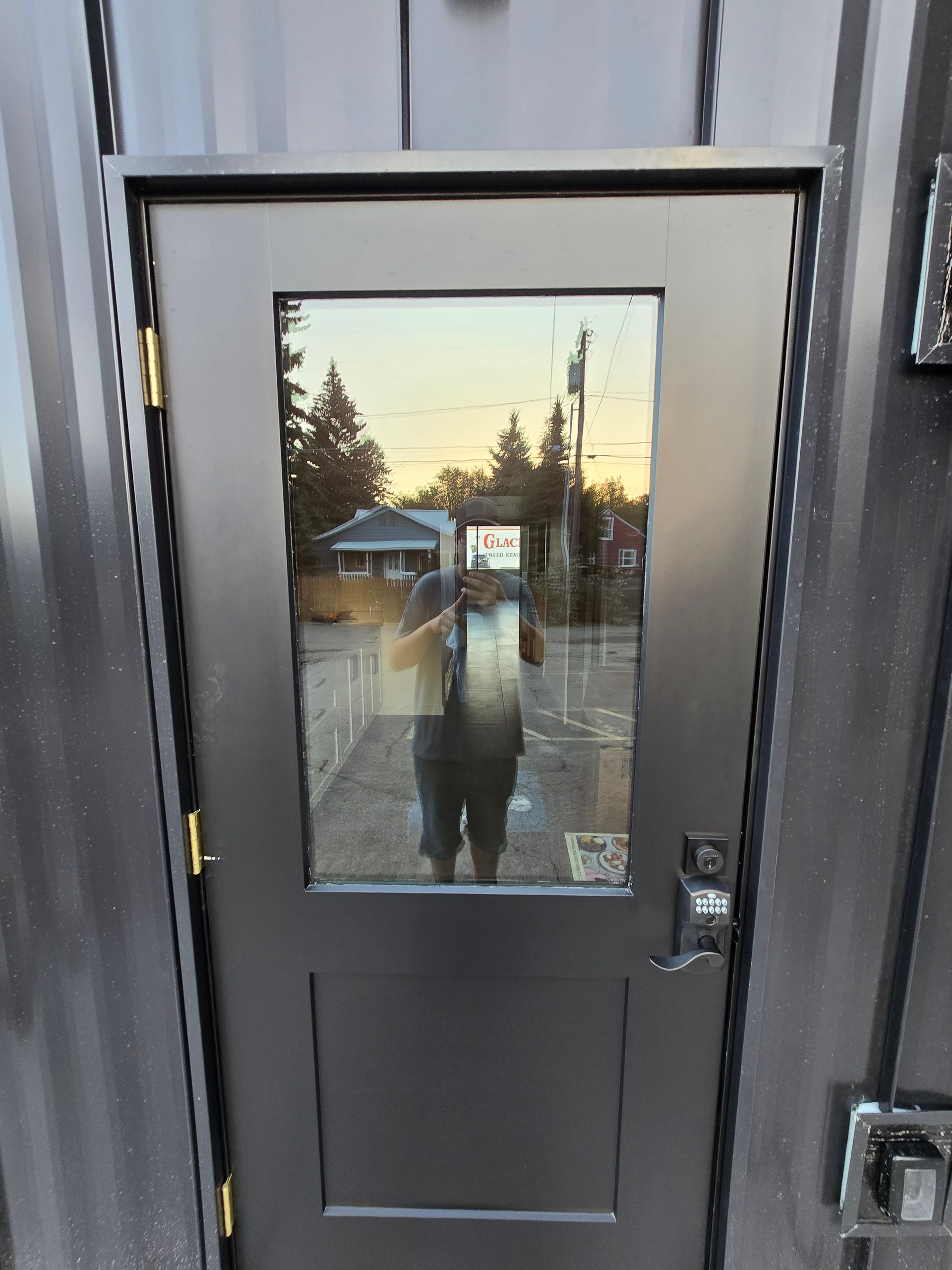 A black door with a rectangular window reflecting an outdoor scene and a person holding a phone to take the photo.