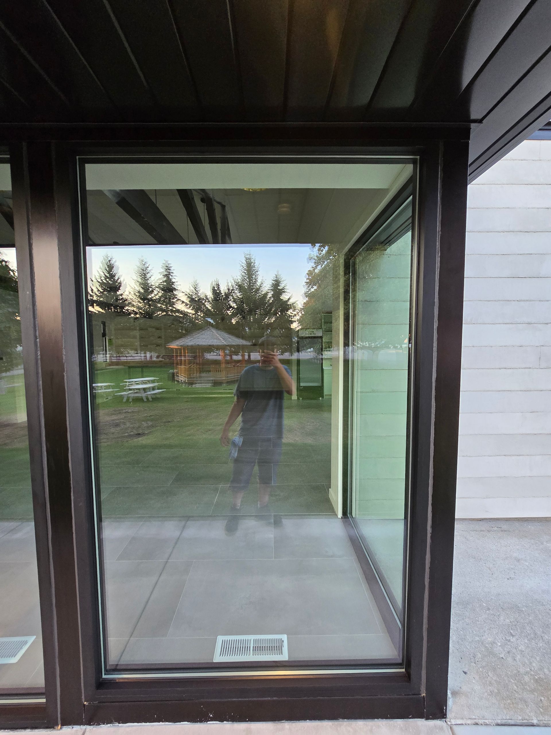 A person reflected in a large glass door leading to a yard with trees and a small wooden structure.