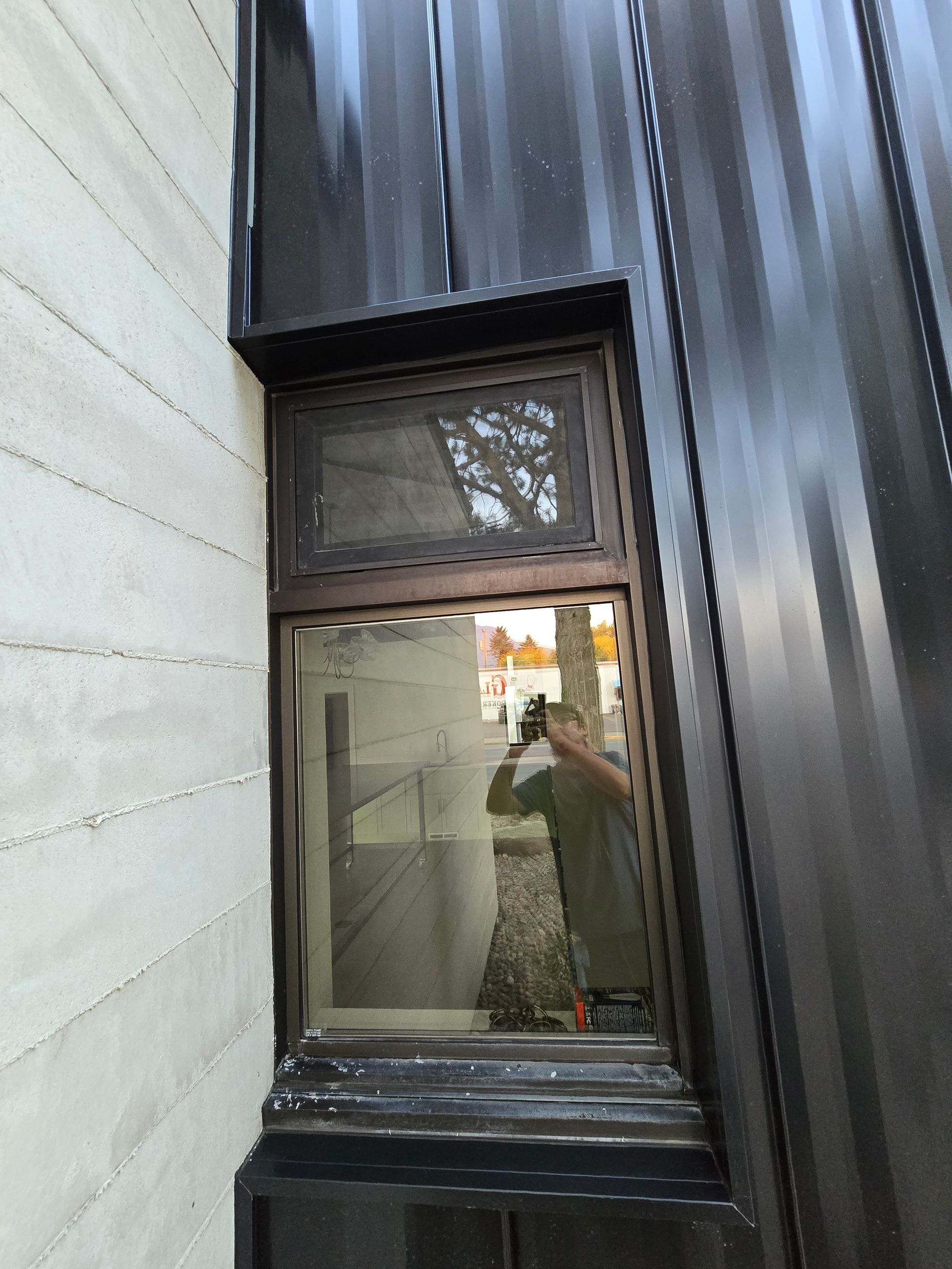 A window set into a dark metal-clad wall, with a partial reflection of a person holding a camera visible in the glass.