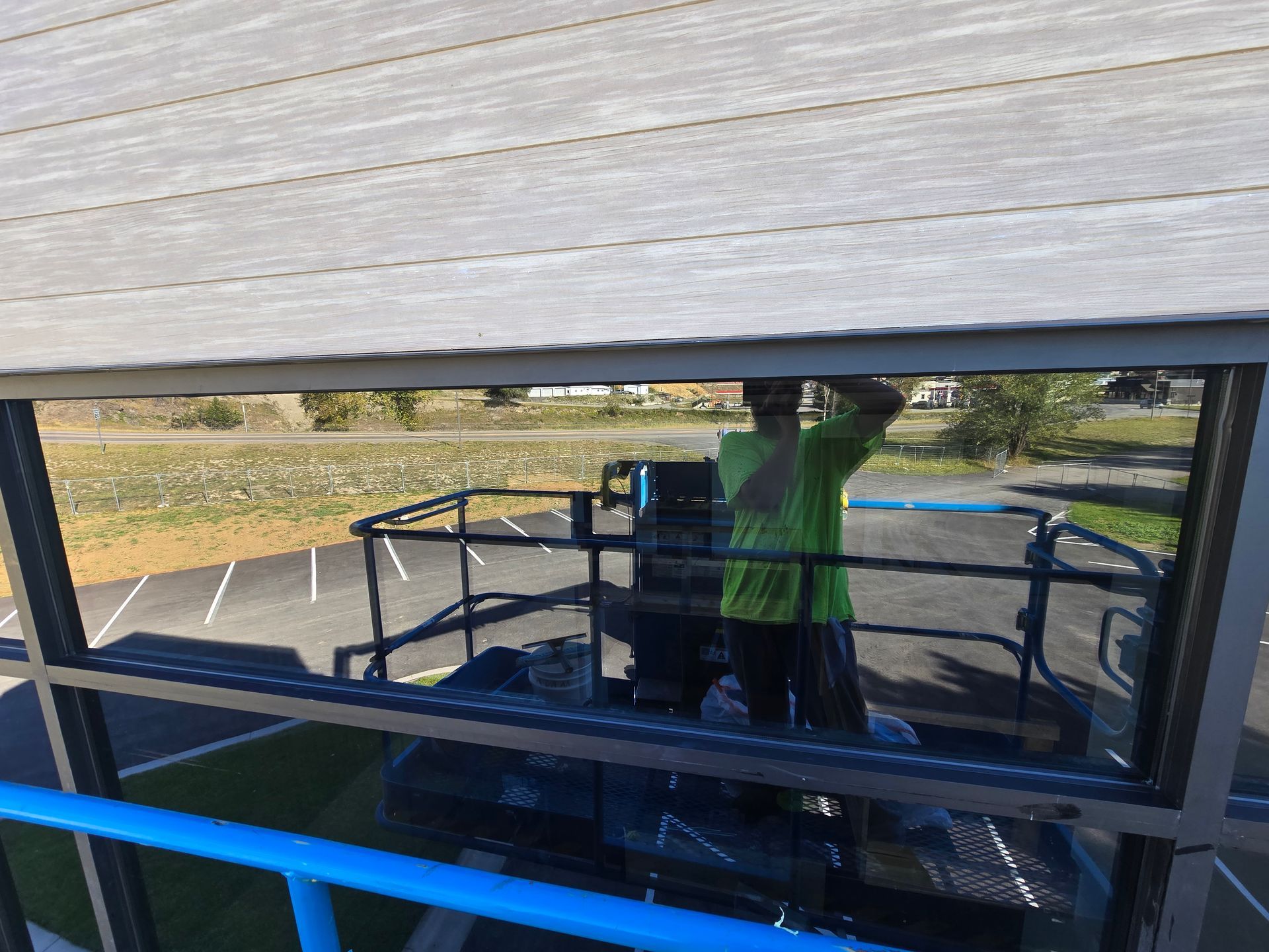 A person in a bright green shirt stands on an elevated lift platform, reflected in a window pane.