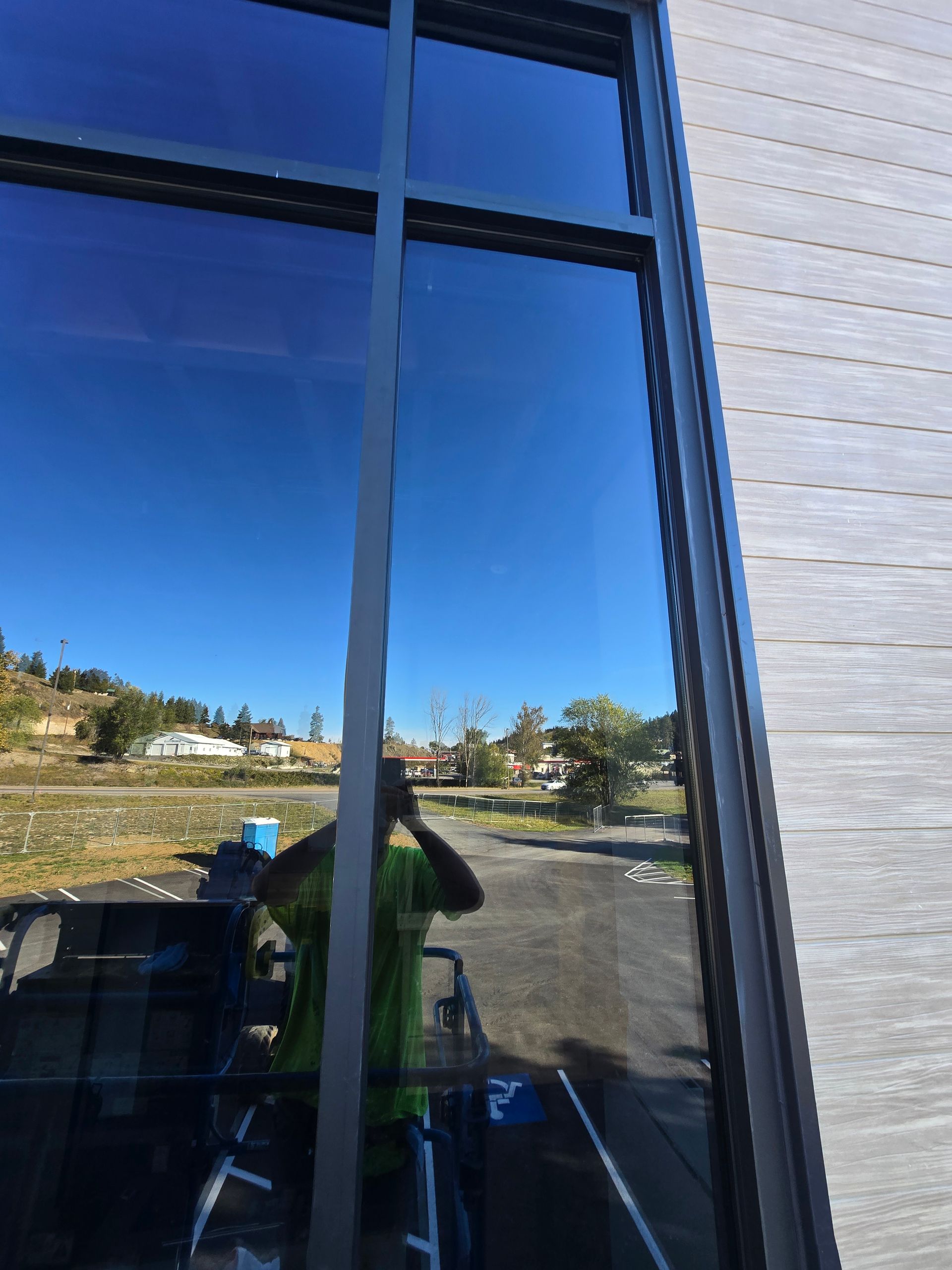 A worker in a bright green shirt reflects in a large glass window of a building with light-colored siding.