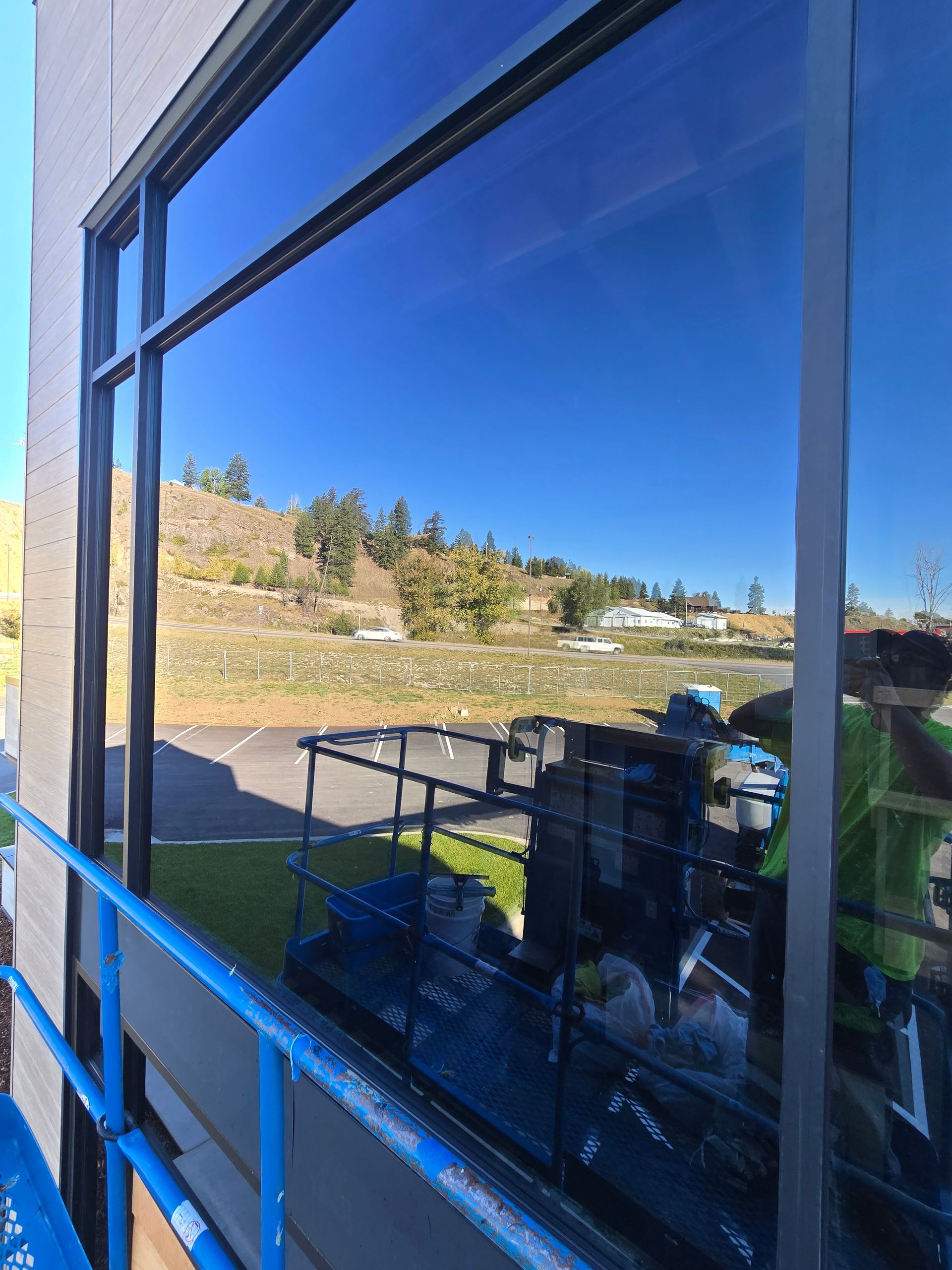 A reflection of a construction lift and worker on the surface of a blue-tinted building window overlooking a grassy hill.