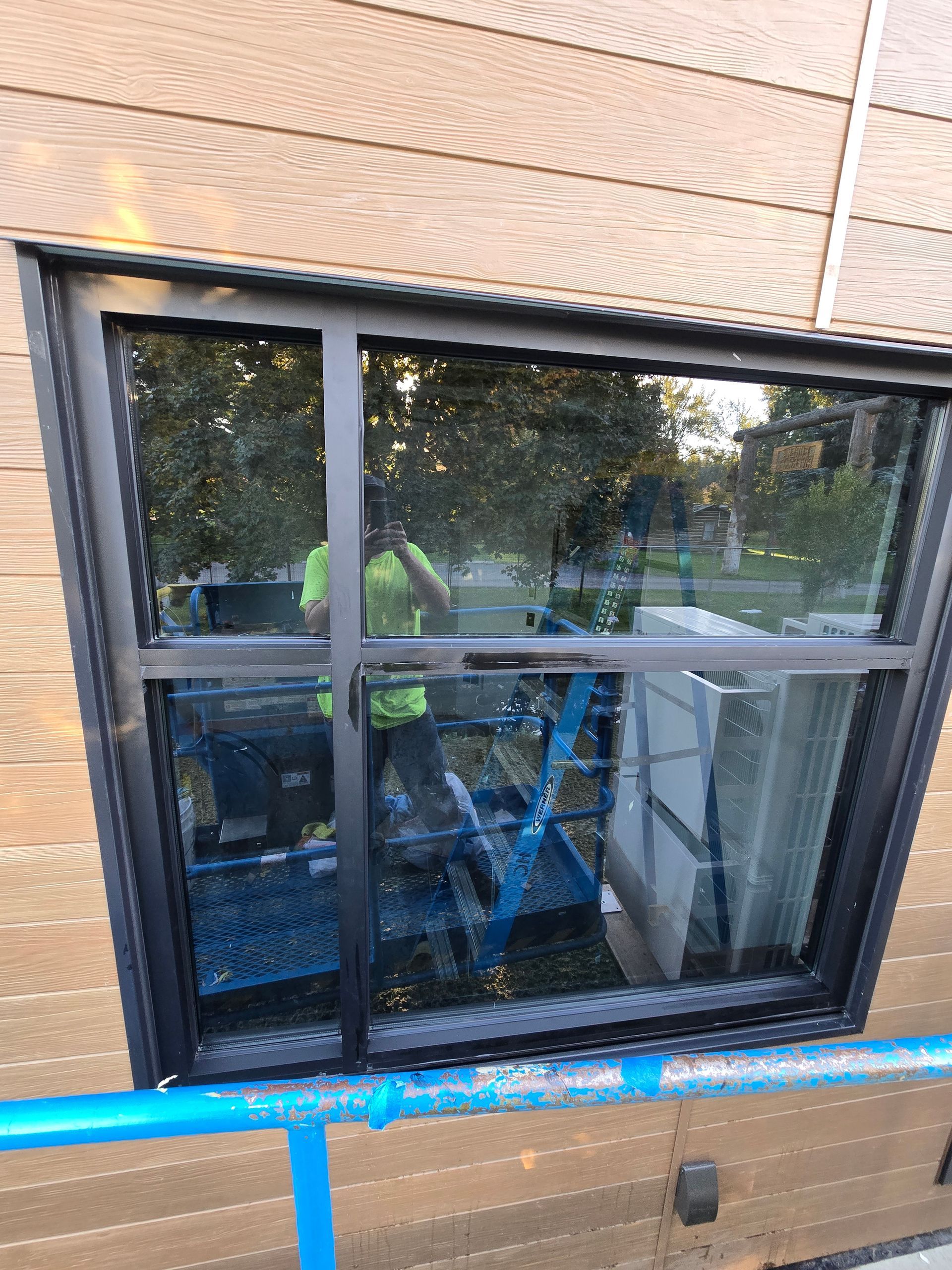 A black-framed window on a wood-sided building reflects a person standing on scaffolding with a blue support bar below.