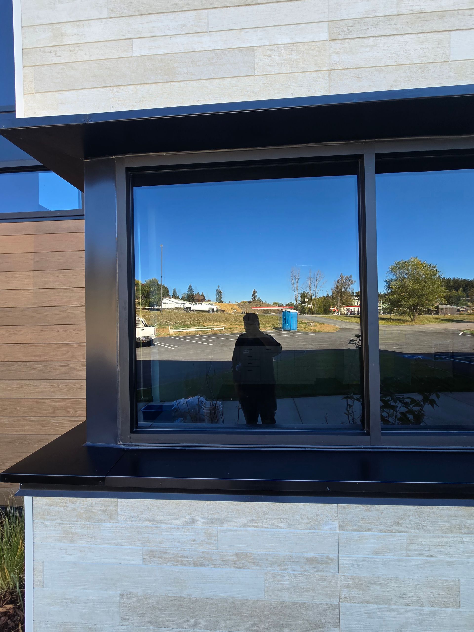 A person stands in front of a reflective glass window on a sunny day, with a building exterior visible behind them.