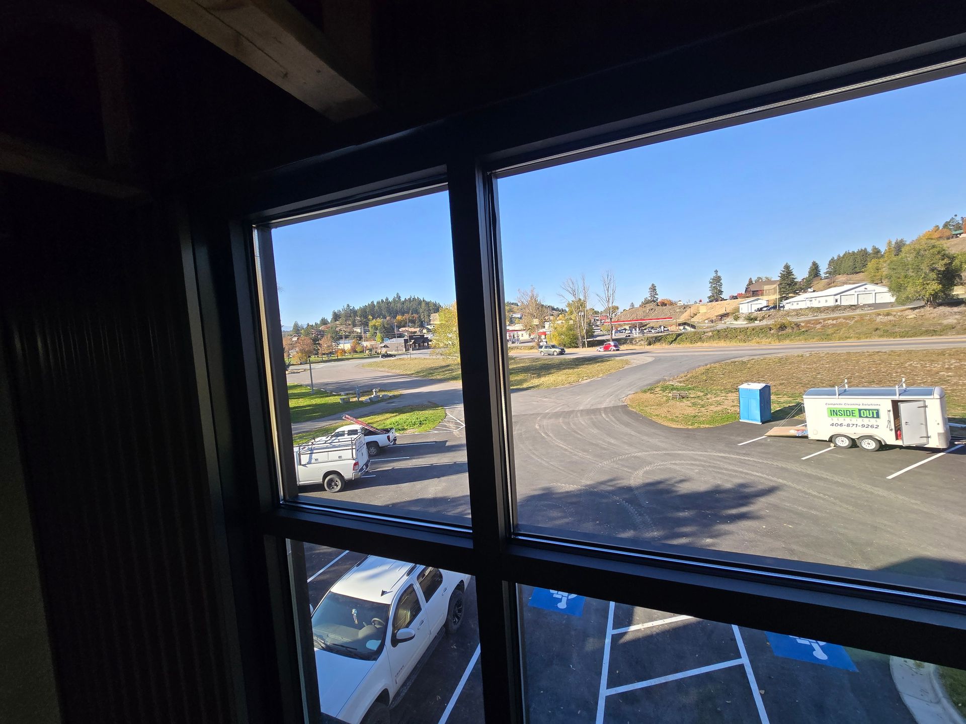View from a window looking out onto a parking lot with several parked cars, a utility trailer, and distant hills.