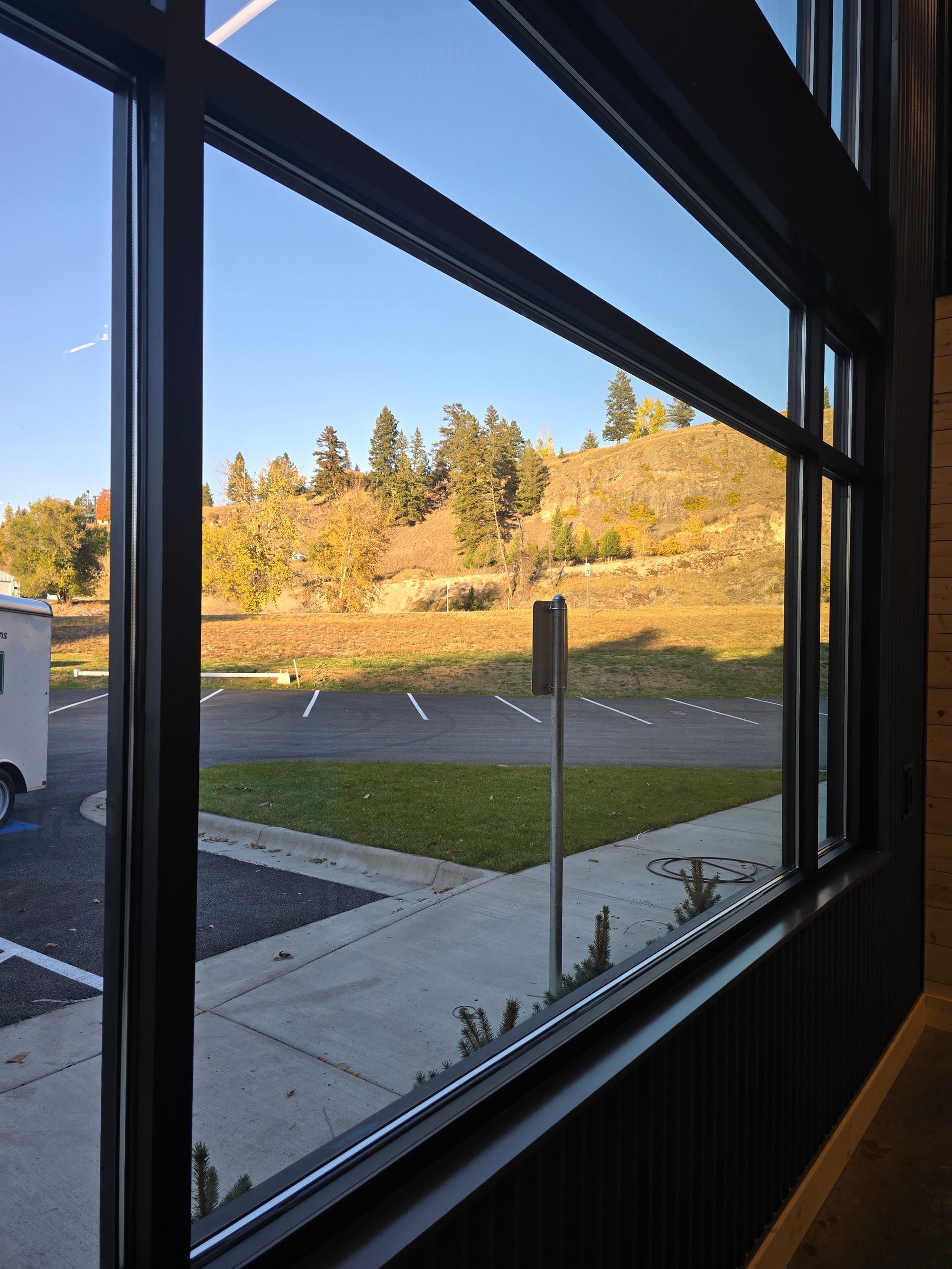 A view through a large window overlooking a parking lot and a grassy hill with trees under a clear blue sky.