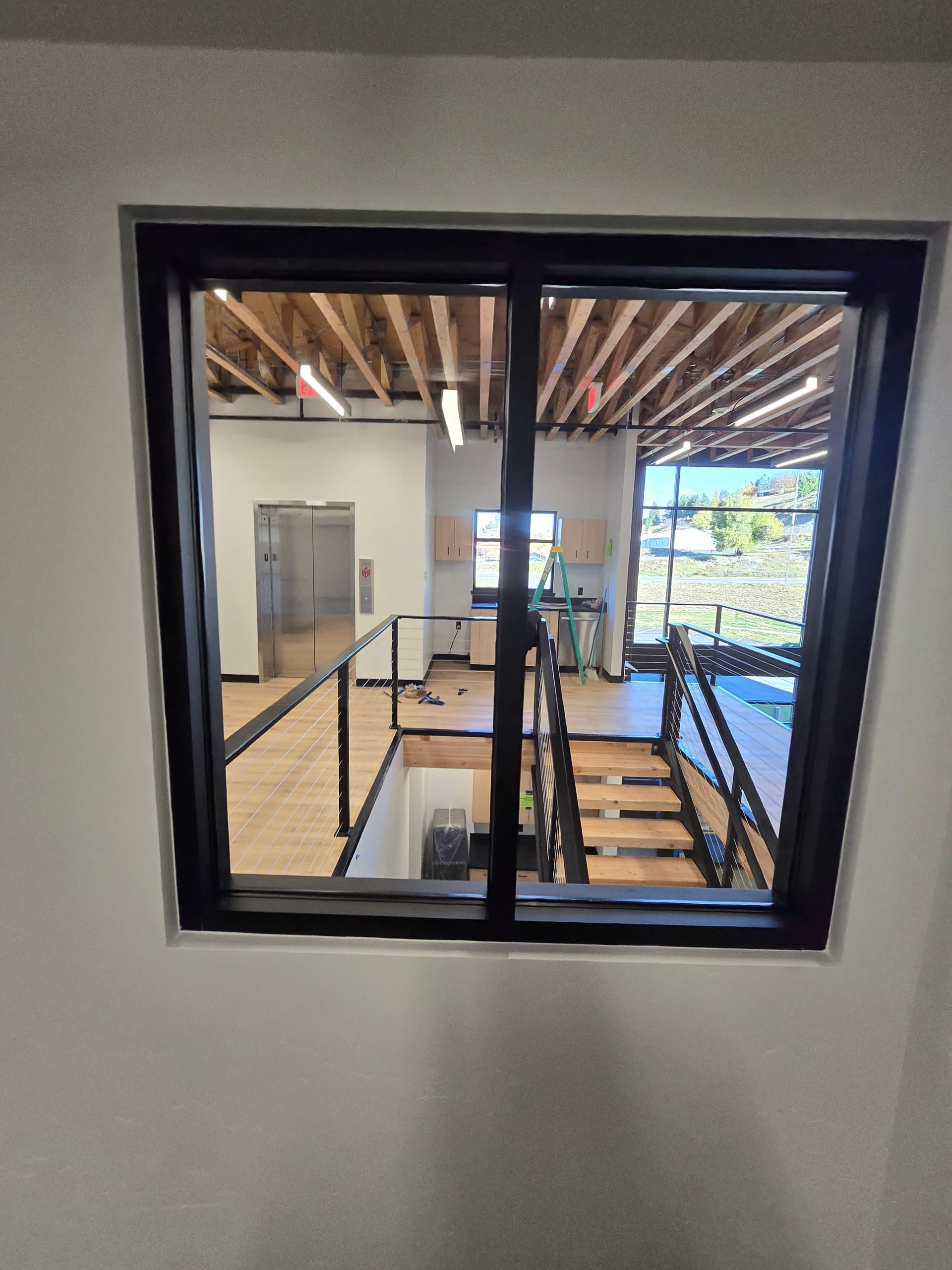 A view through a black-framed window showing an interior staircase, a landing with railings, and an open, unfinished space.