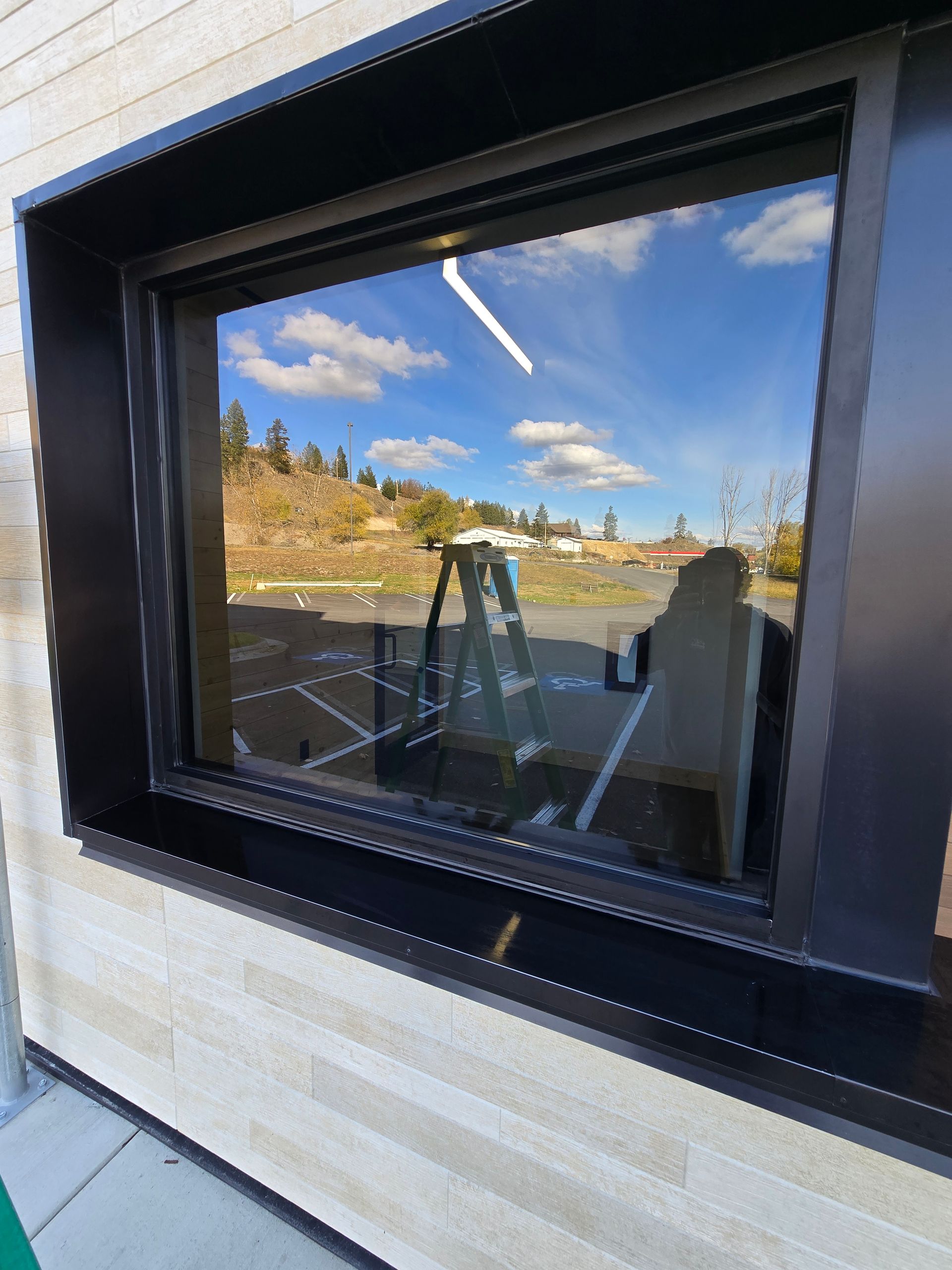A black-framed window on a beige brick building reflects the sky, a ladder, and a parking lot.