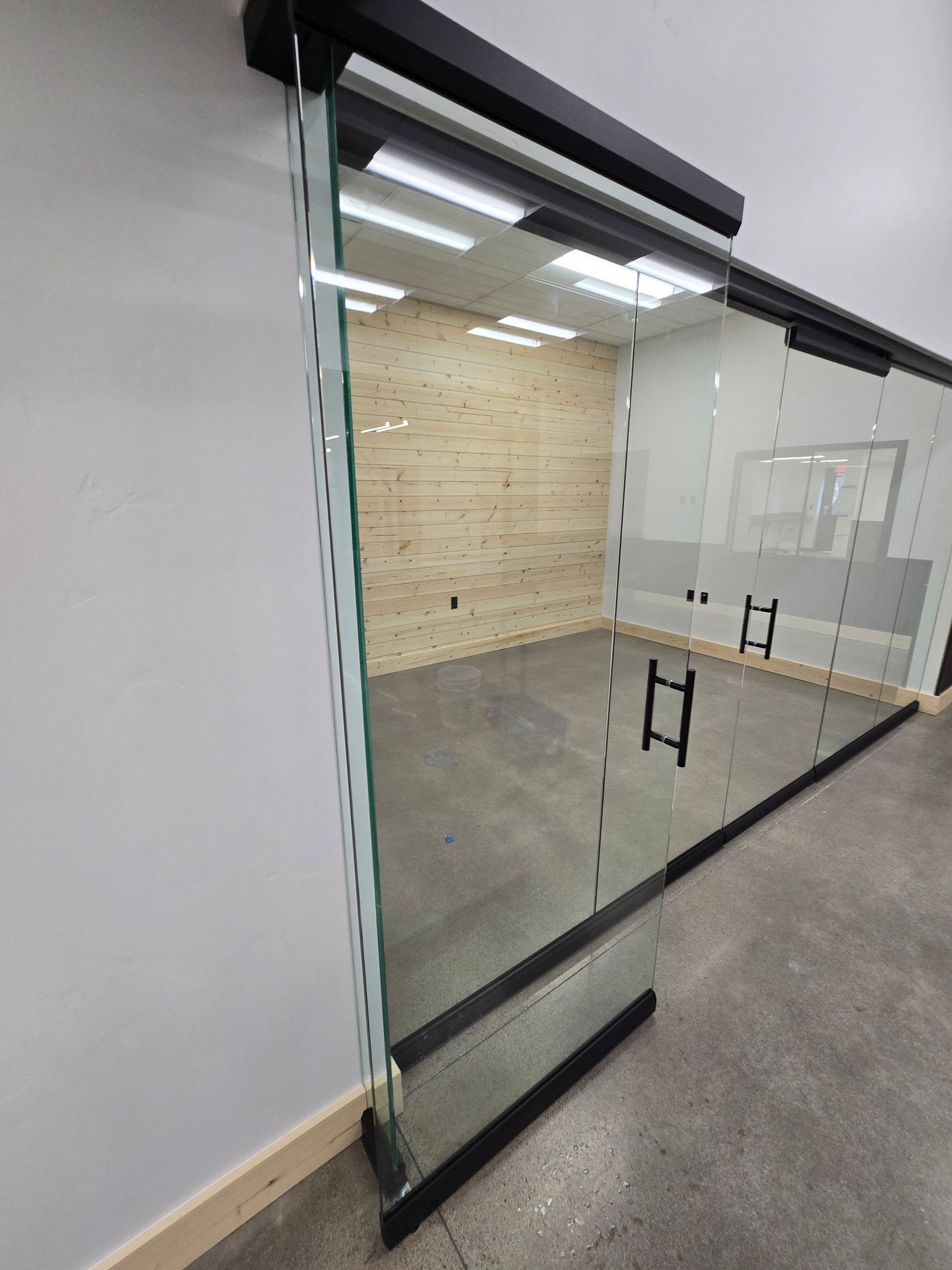 A modern glass office partition with a sliding door and black metal frame, opening into an empty room with wood walls.