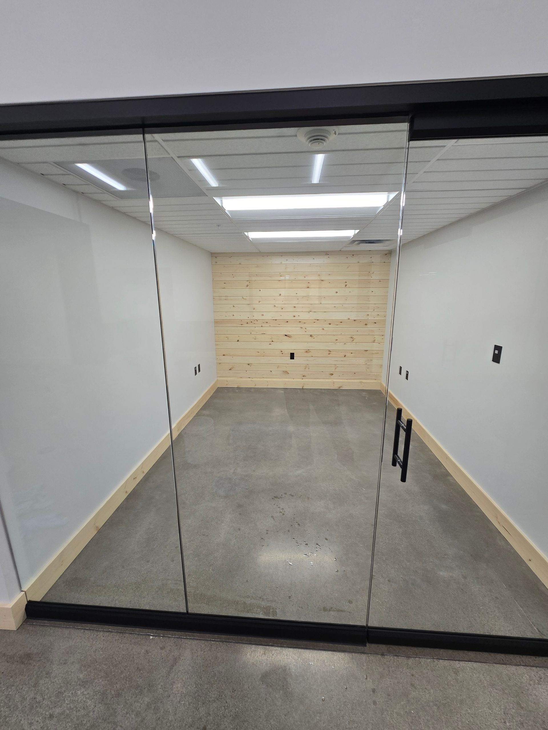 A small, modern office with glass entry doors, grey concrete floors, white walls, and a light-wood plank back wall.